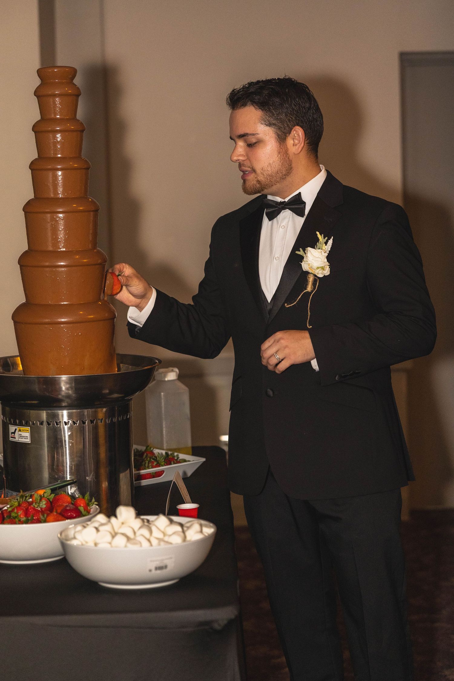 Groom in black tuxedo dipping strawberry into chocolate fountain at elegant wedding reception.