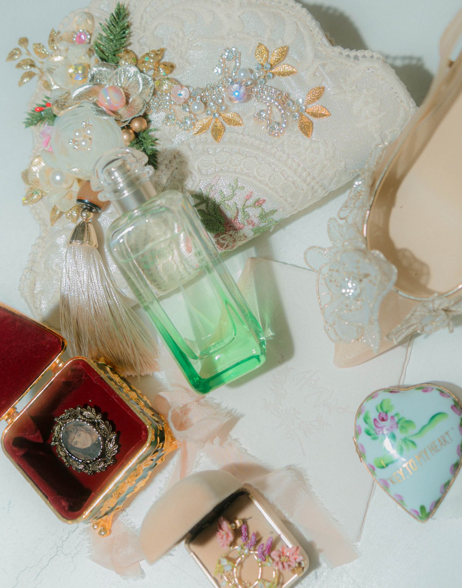 Flat lay of vintage bridal accessories including a green perfume bottle, floral headpiece, ring box, and lace items.