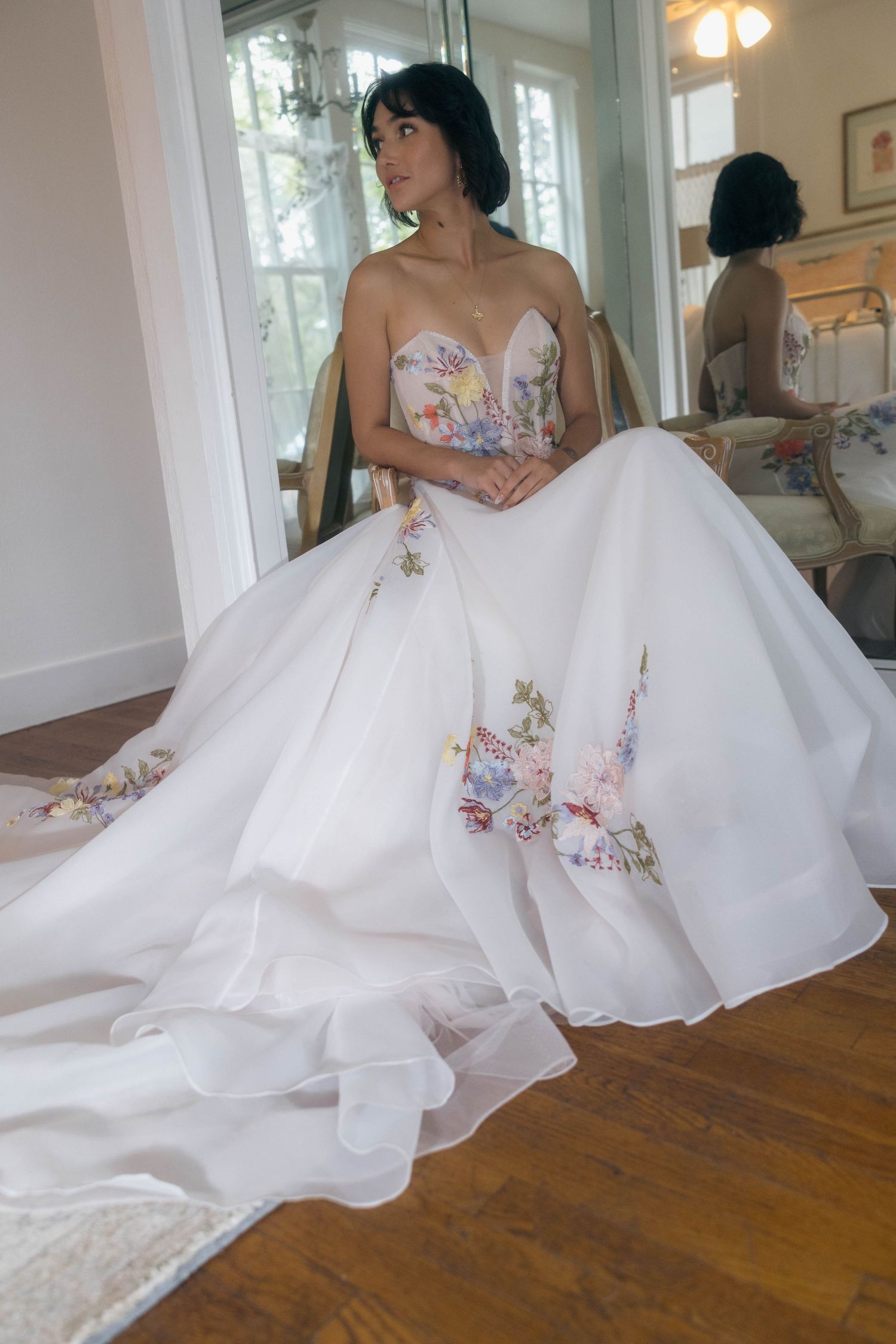 A bride sits elegantly in a floral embroidered strapless ball gown with a long train in a sunlit room.