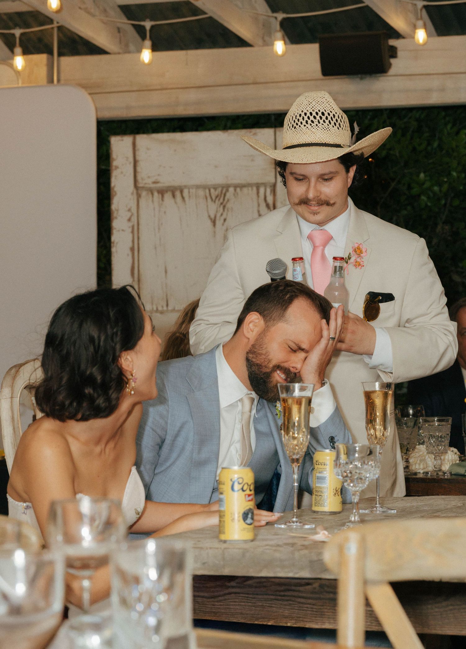 Groomsman in cowboy hat playfully blessing groom at rustic outdoor wedding reception dinner.