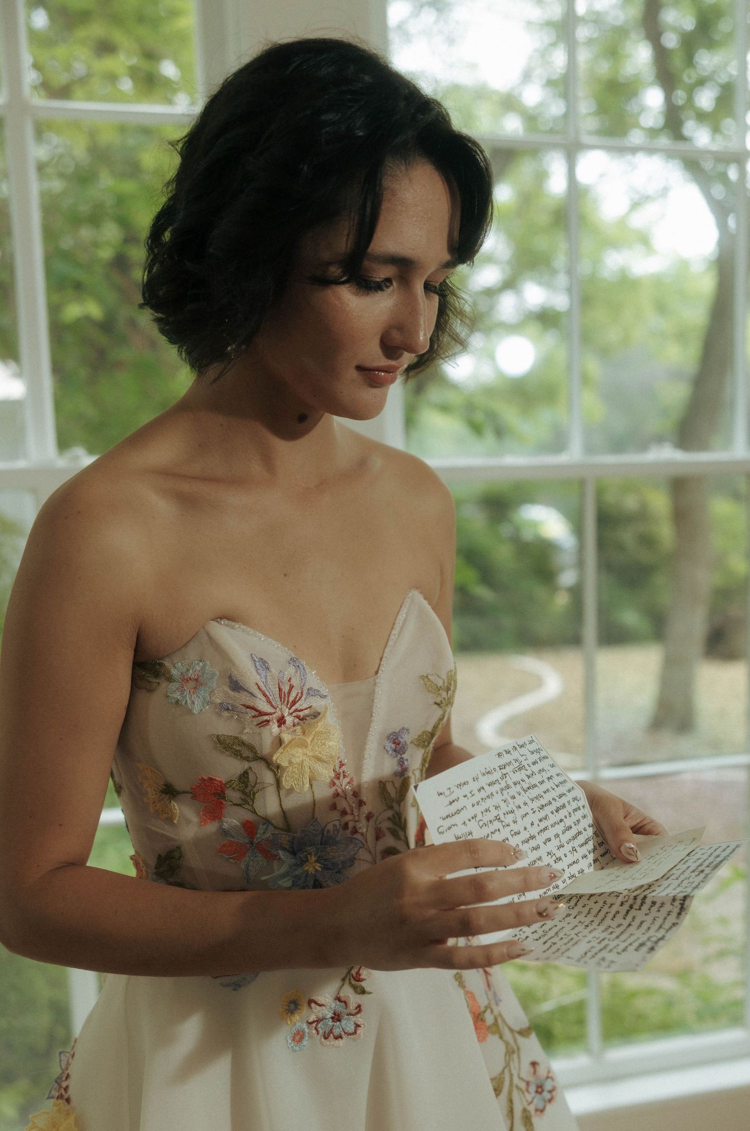 Bride in floral strapless gown reading handwritten letter by sunlit window indoors.

New Braunfels Brides