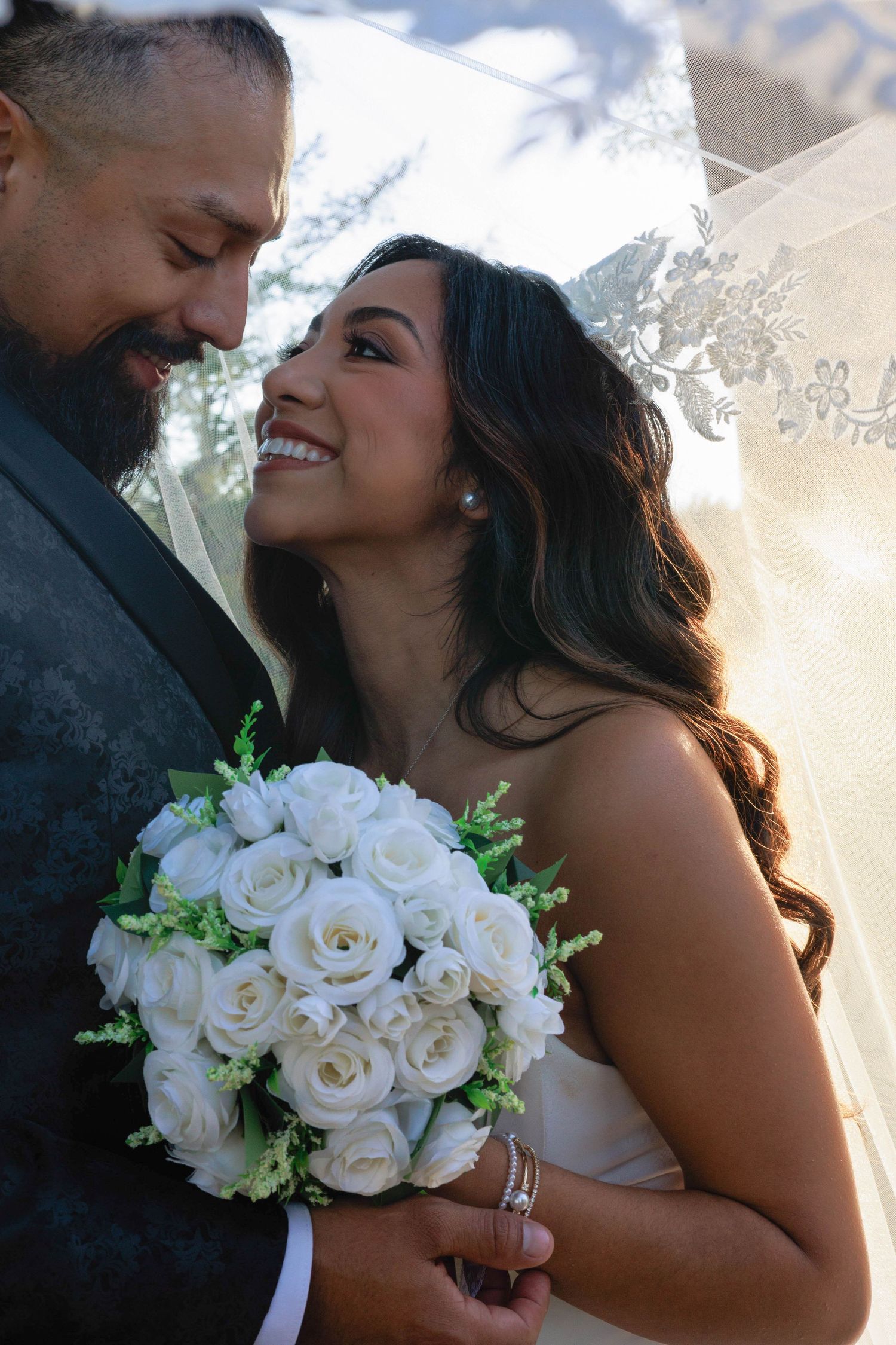 Joyful bride holding white rose bouquet smiles up at bearded groom in intimate outdoor portrait.