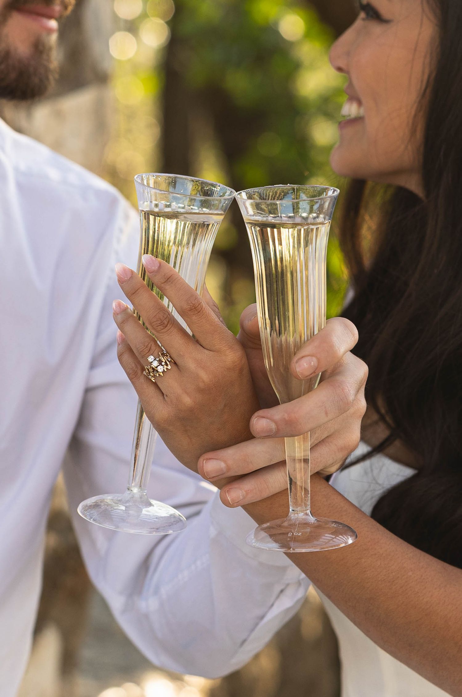 Smiling bride and groom clinking champagne glasses together outdoors in soft natural light.