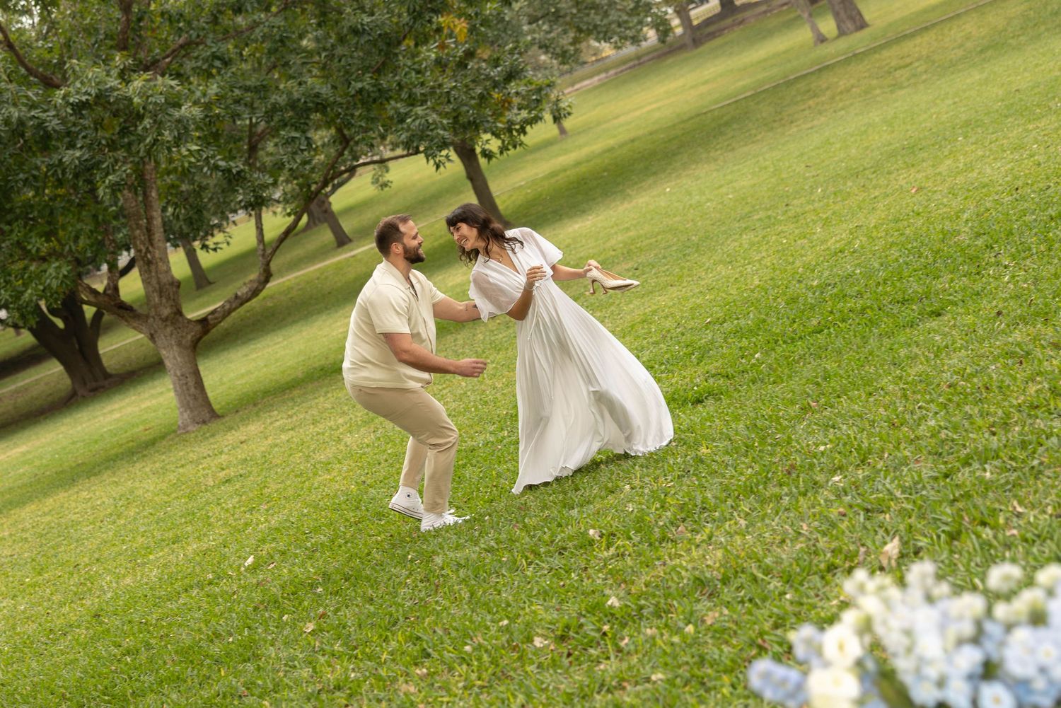 Couple dancing playfully on green grass in a park, woman in white dress, man in beige outfit.