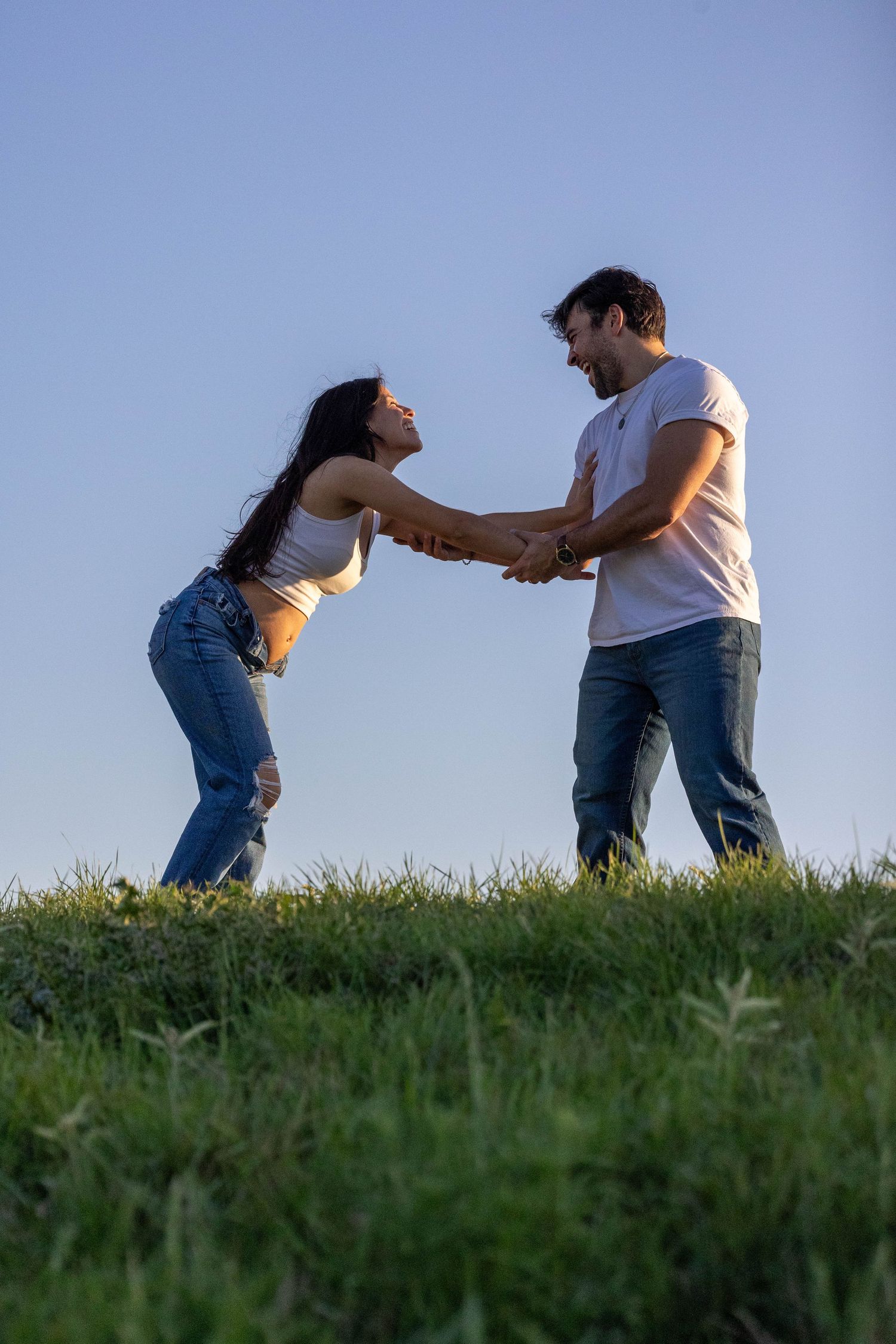 A laughing couple holds hands and leans back playfully on a grassy hill under a clear blue sky.