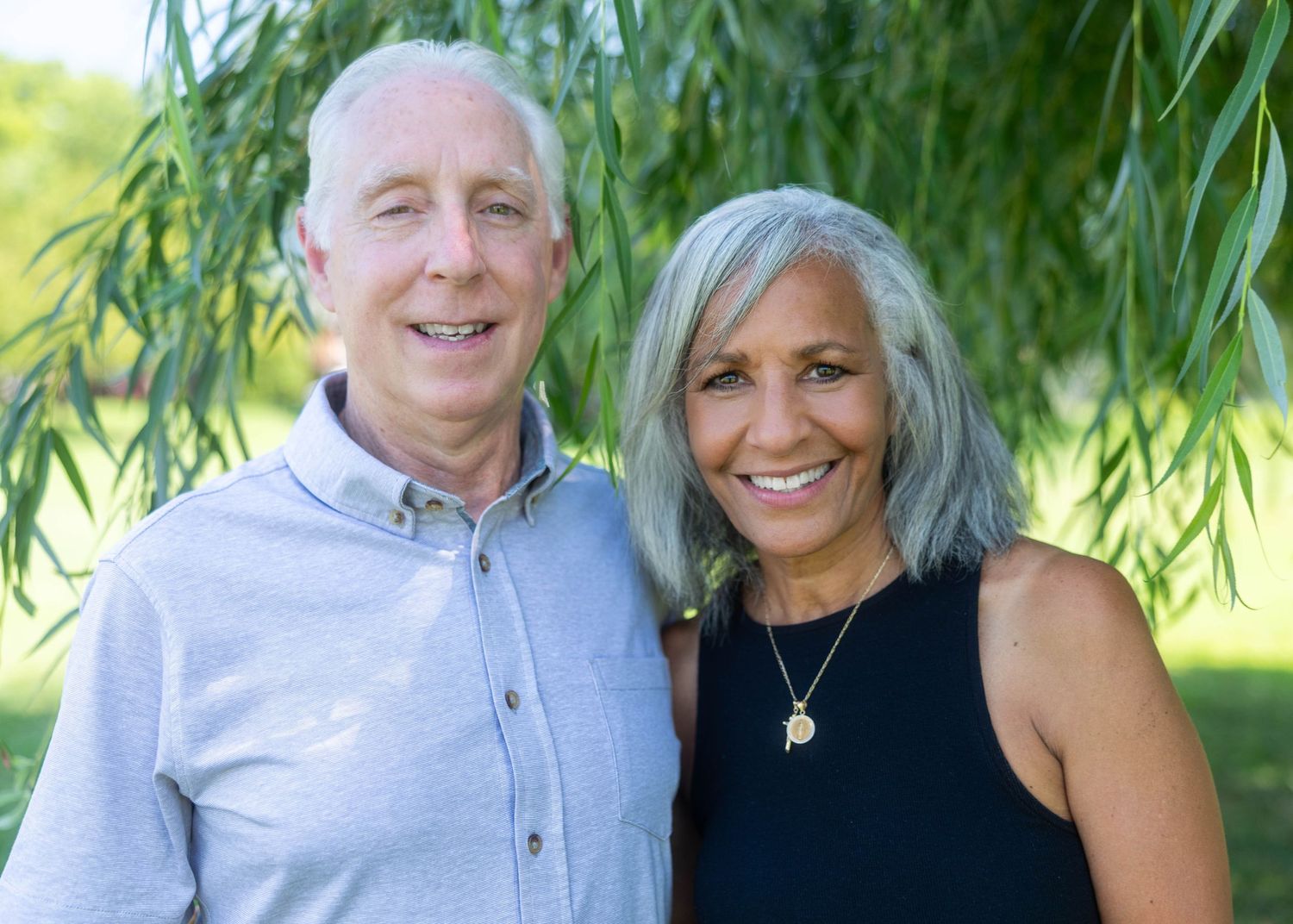 Smiling older couple posing outdoors under a willow tree in a lush green park setting.