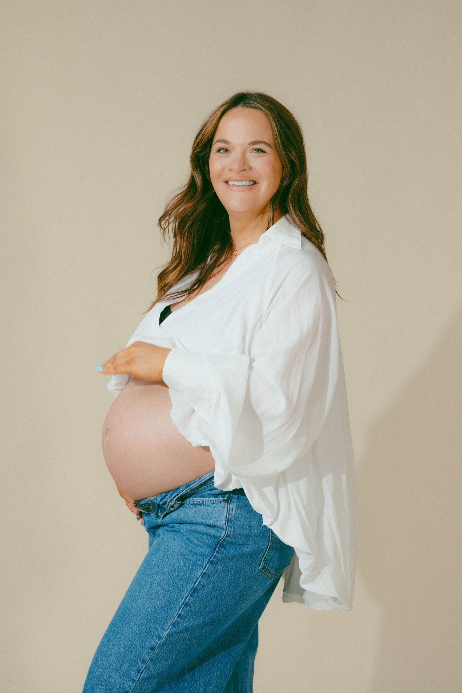Smiling pregnant woman in white shirt and jeans cradles bare baby bump in bright studio maternity portrait.
