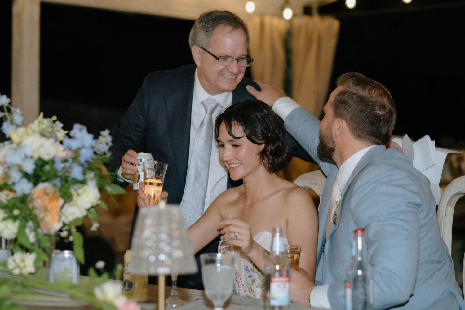 Older gentleman leans in to speak with bride and groom seated at floral-decorated wedding reception dinner table.
