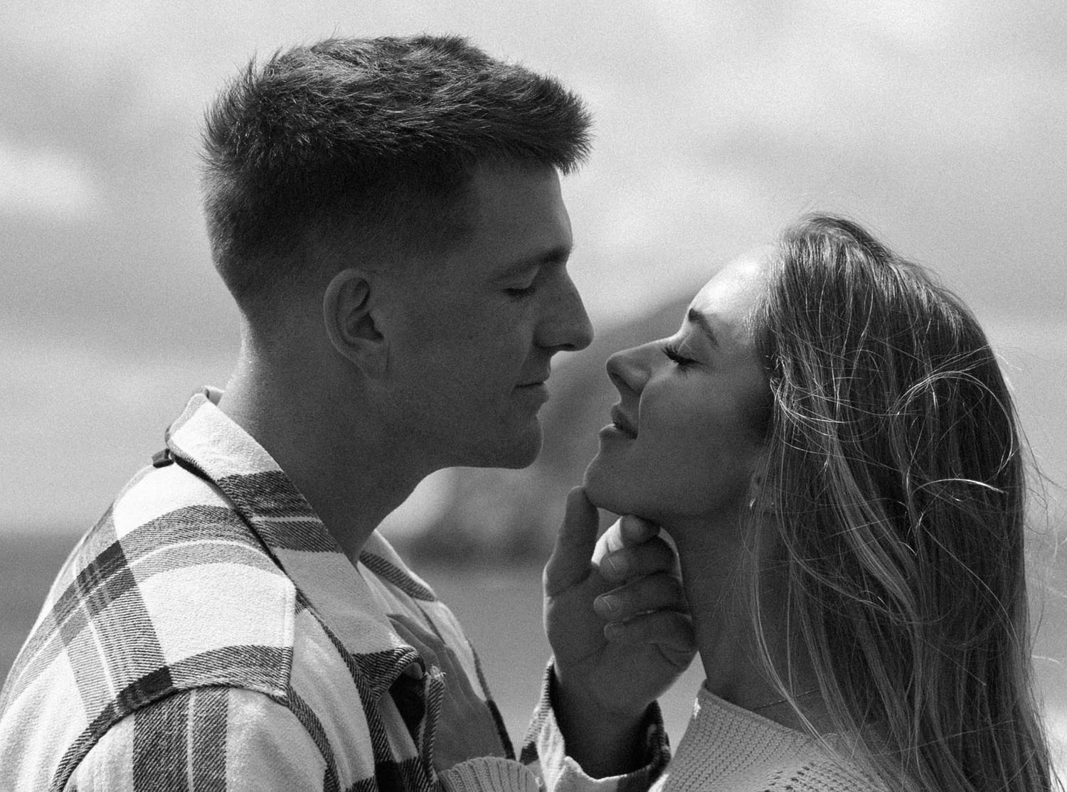 Romantic black and white close-up portrait of couple nearly kissing under cloudy sky outdoors.