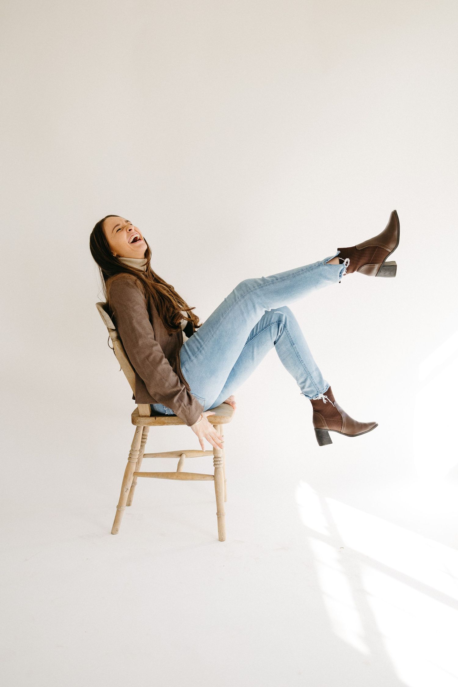 Joyful person wearing brown jacket and light jeans kicks up brown boots while sitting on wooden chair against white wall.
