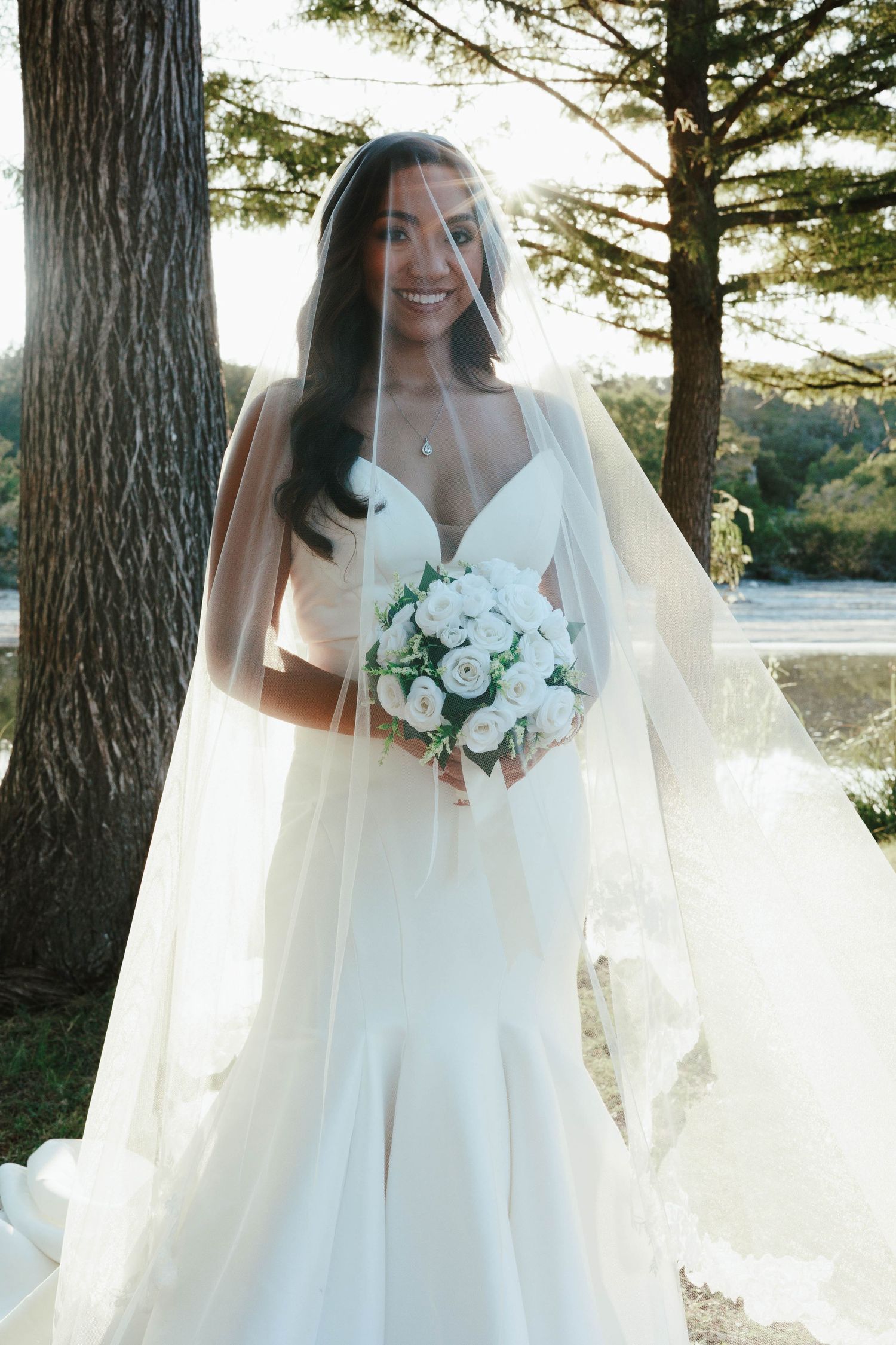 Radiant bride in mermaid wedding dress and cathedral veil holds white bouquet in sunlit garden.