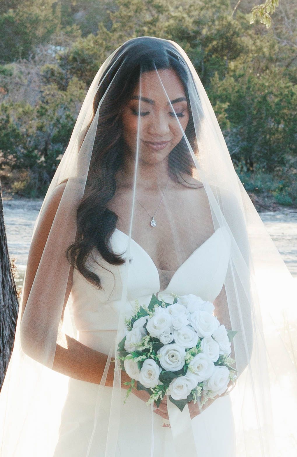 Beautiful bride wearing a white veil and gown holding a bouquet of white roses in an outdoor setting.