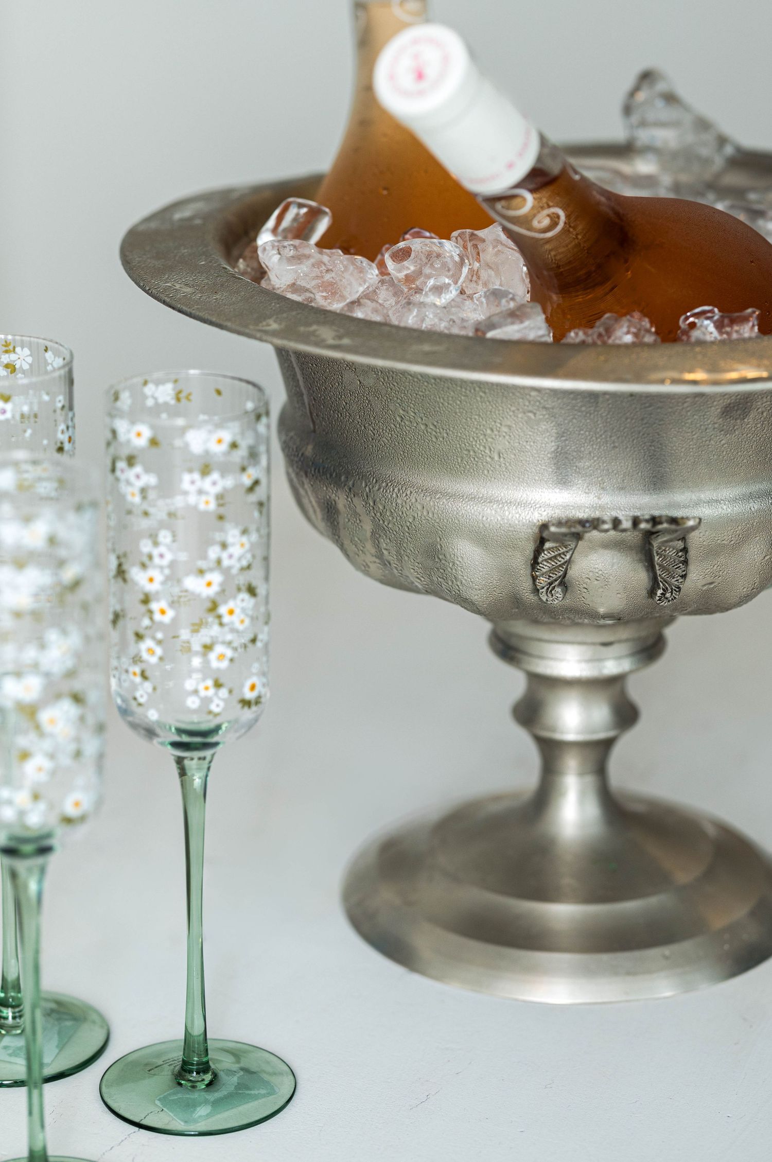 Elegant champagne bottle chilling in a silver ice bucket beside two daisy-painted crystal flute glasses on a white surface.