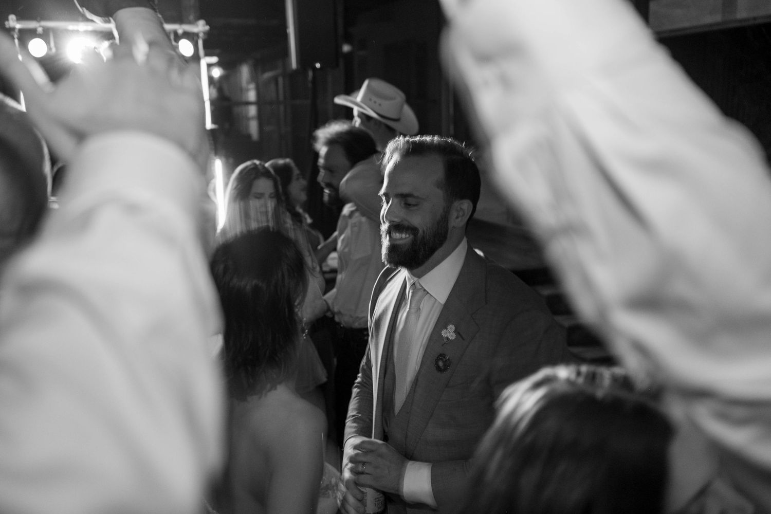 Smiling groomsman in a suit celebrates on the dance floor surrounded by wedding guests.