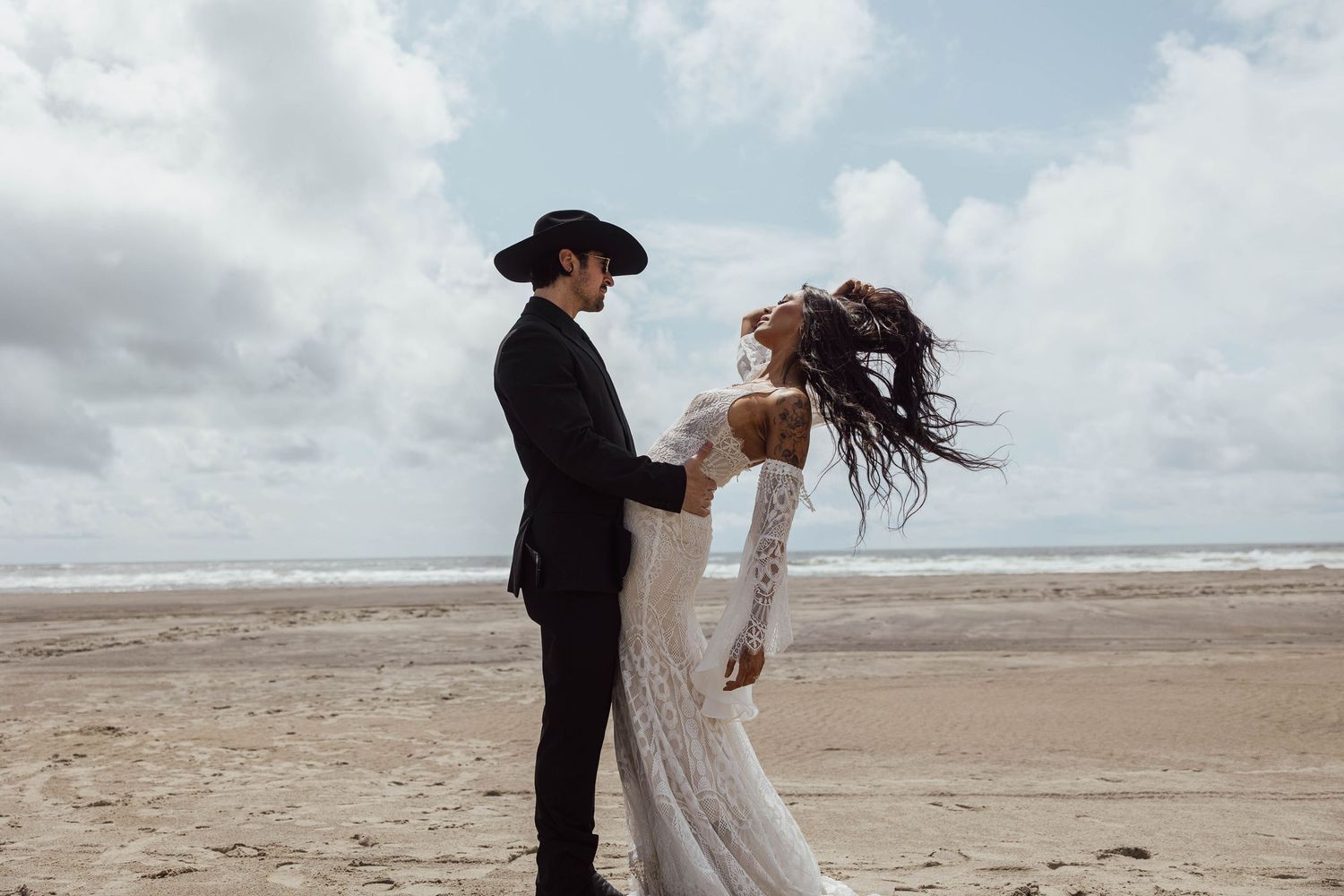 Couple in wedding attire dancing romantically on a sandy beach with cloudy skies.