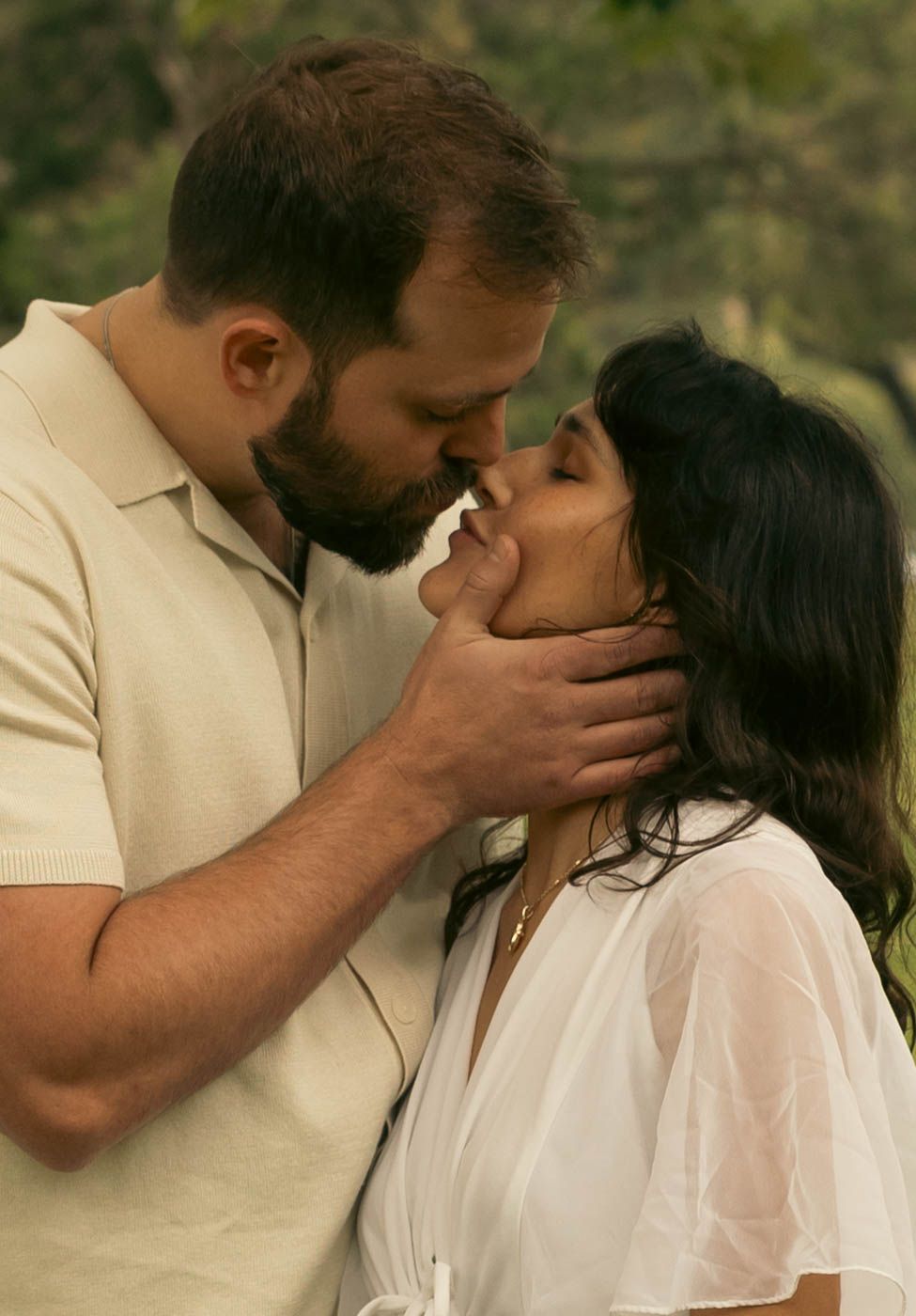 Bearded man tenderly cupping woman's face for a kiss outdoors in a lush green setting.