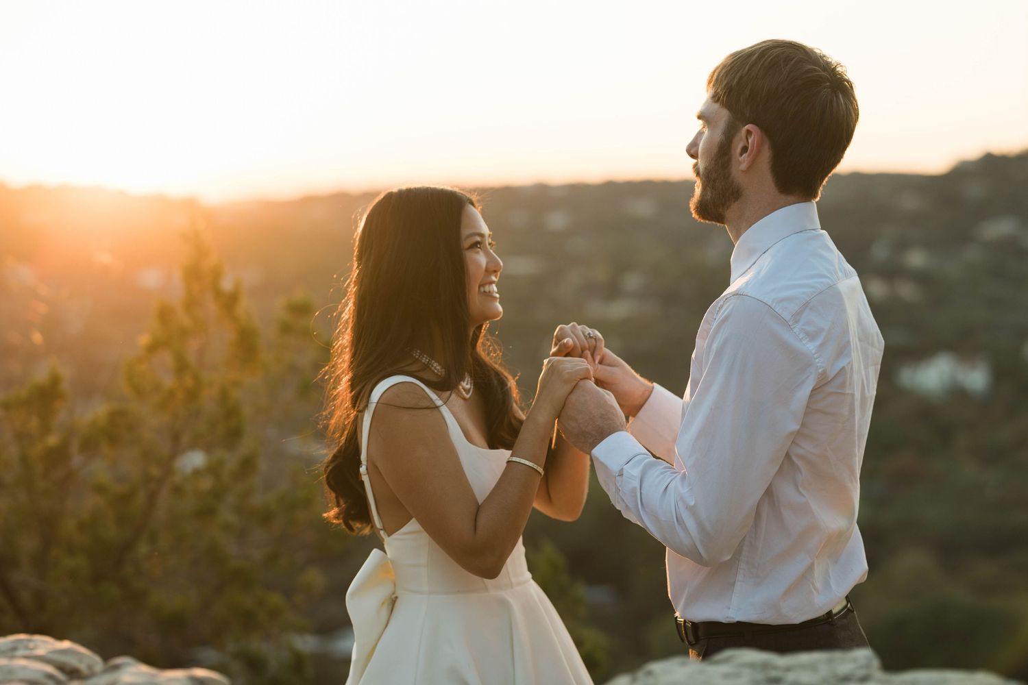 Couple holding hands at sunset with scenic hilltop landscape in background.