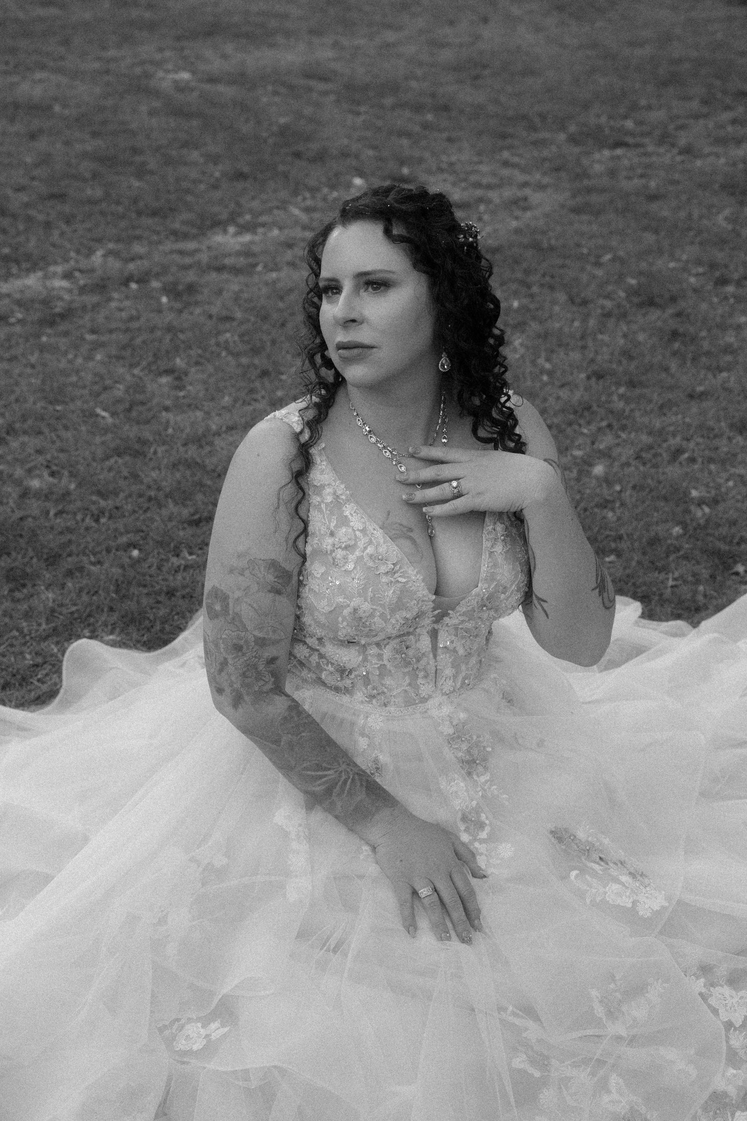 Black and white portrait of tattooed bride in elegant lace wedding gown sitting gracefully on green grass.