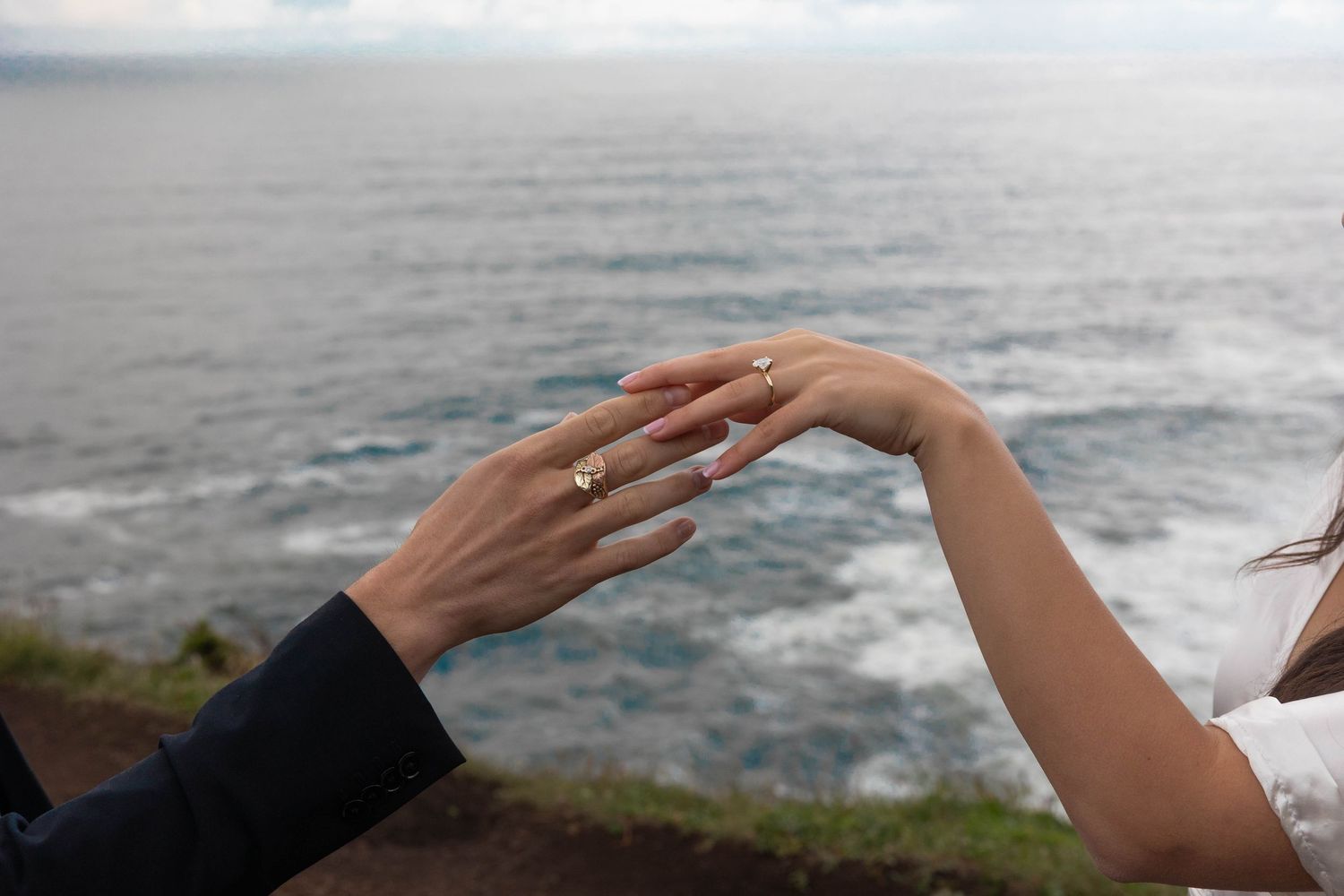 Bride and groom touch hands displaying gold wedding rings with ocean backdrop at coastal overlook.