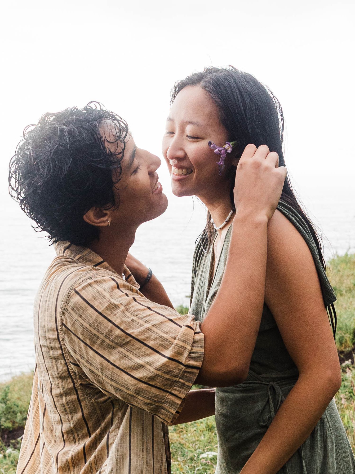 Happy couple almost kissing outdoors by the sea, woman with purple flowers tucked behind her ear laughing.