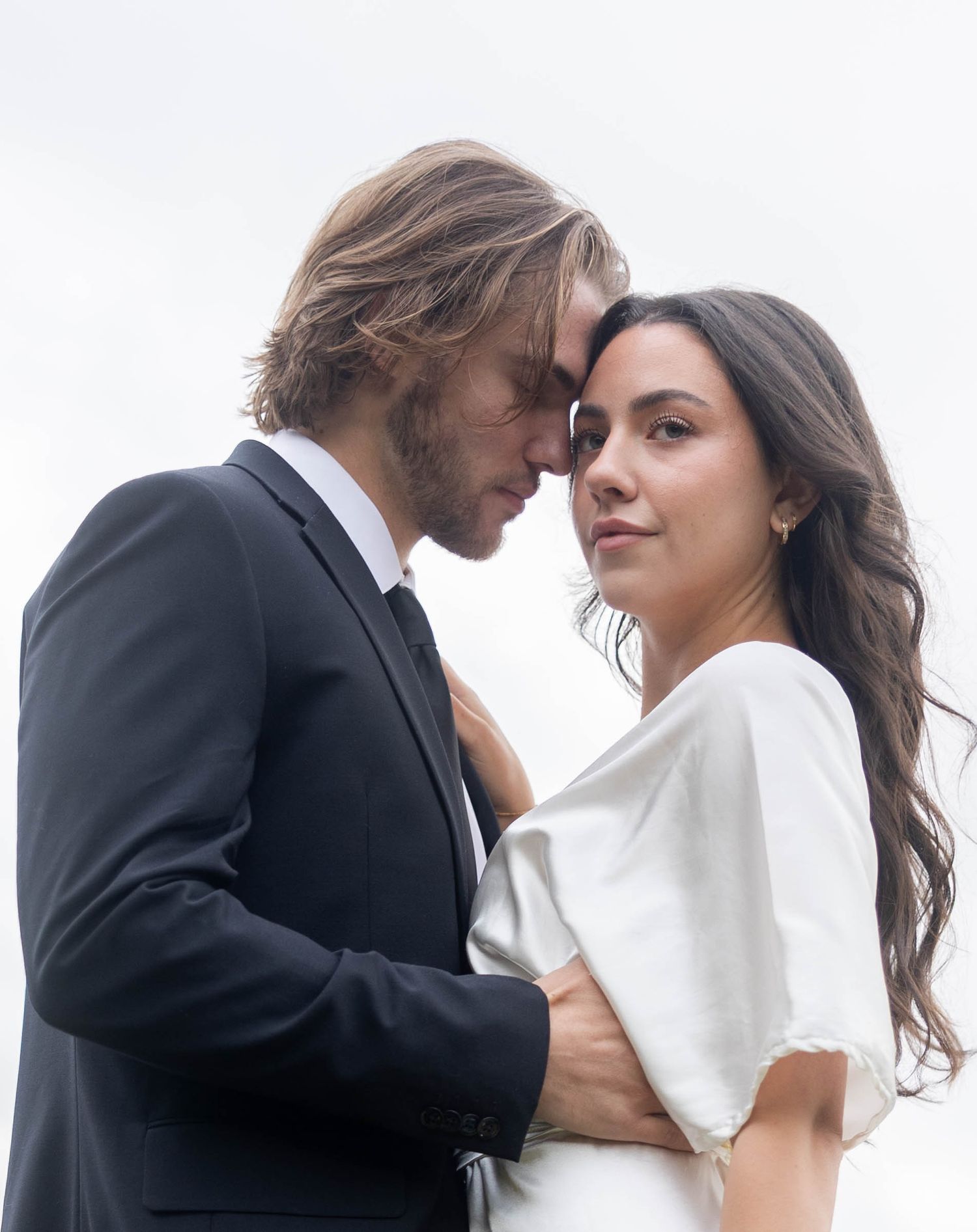 Elegant couple in formal attire touching foreheads intimately against a bright white background.