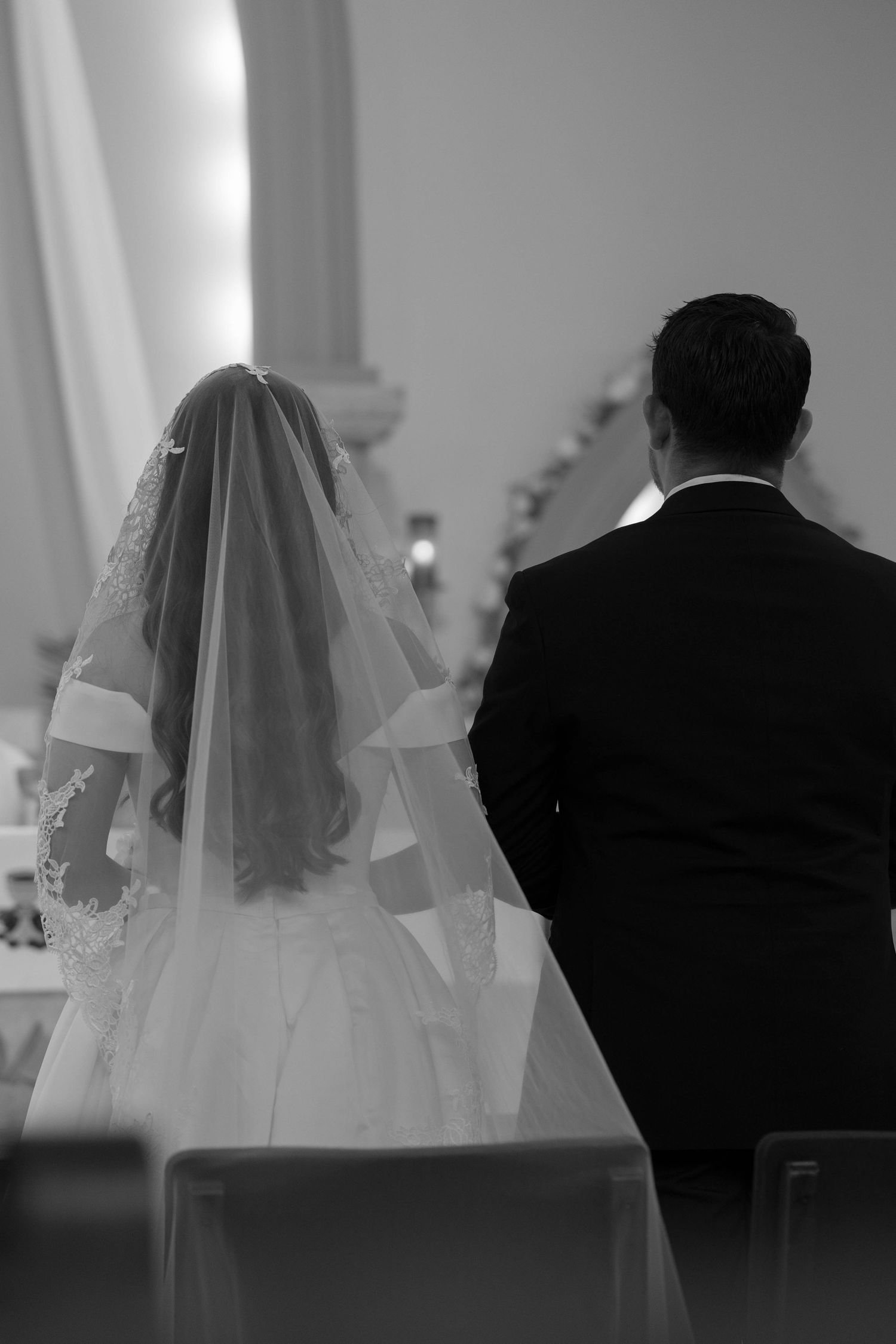 Black and white rear view of bride in lace veil and groom in tuxedo seated during church ceremony.