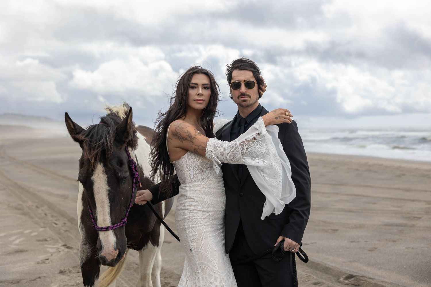 Bride in lace gown and groom standing with a horse on a windy beach.
