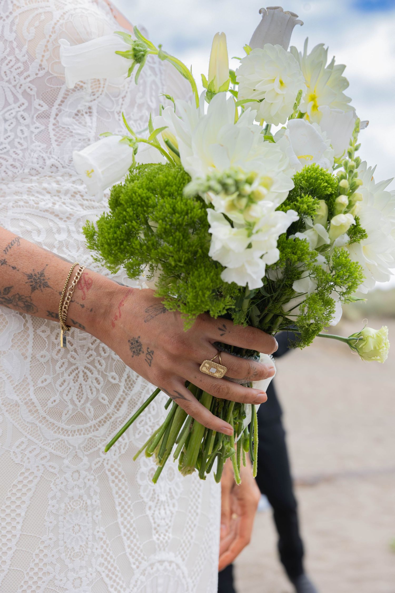 A delicate white floral bouquet with green accents is held by a hand adorned with rings and tattoos against a lace backdrop.