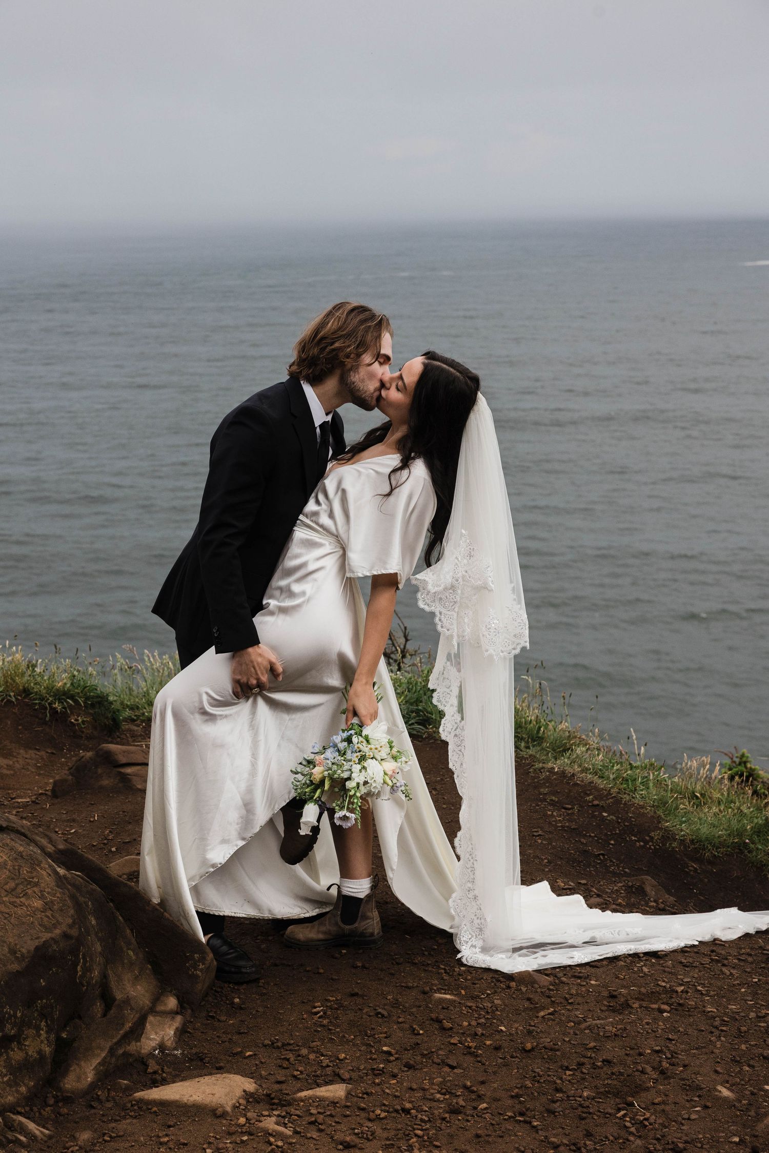 Bride and groom share a passionate kiss on a rocky coastal overlook, bride in satin gown holding white bouquet.