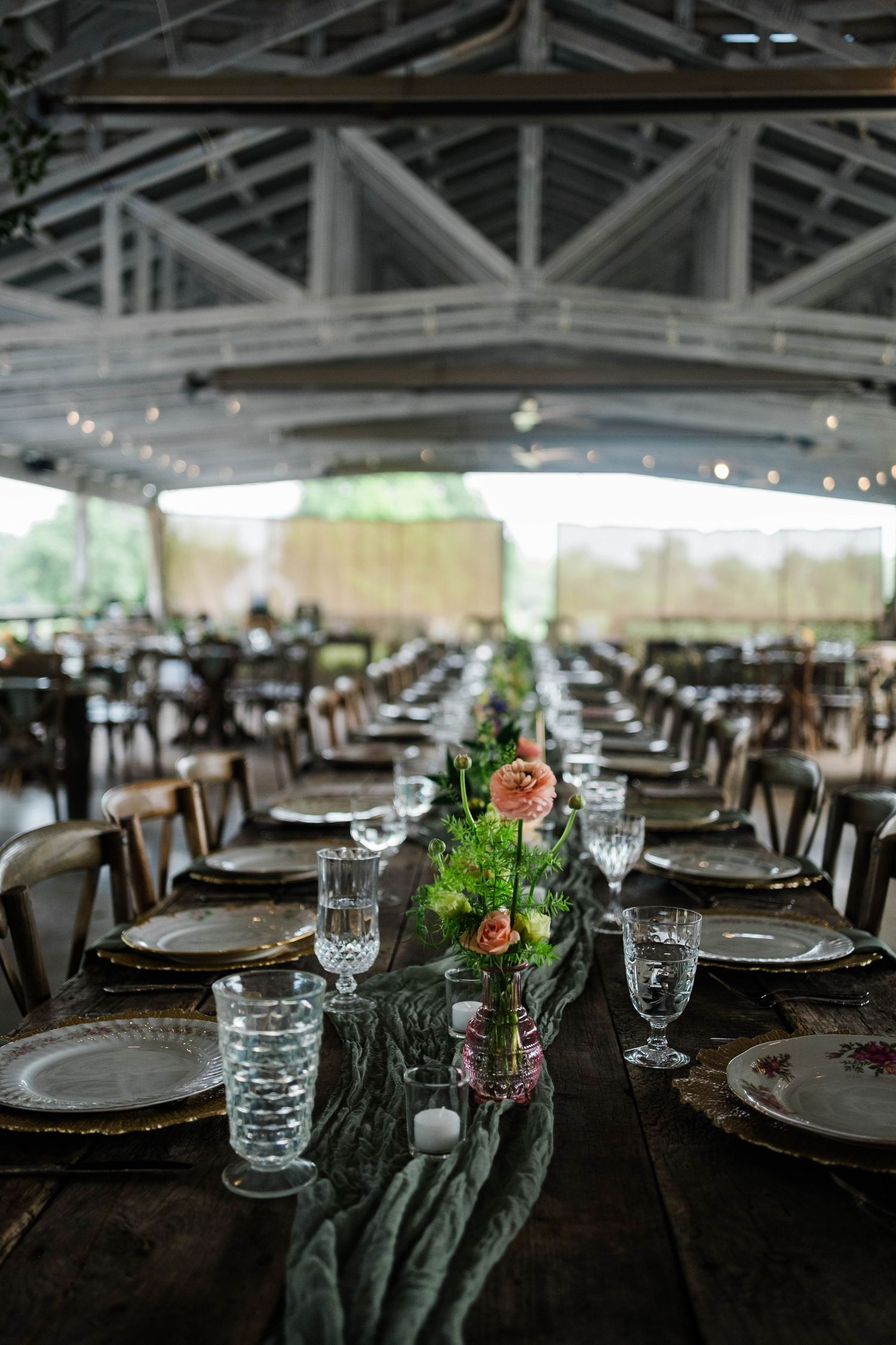 Long rustic farm tables beautifully set with green runners, floral centerpieces, and glassware under a tent.