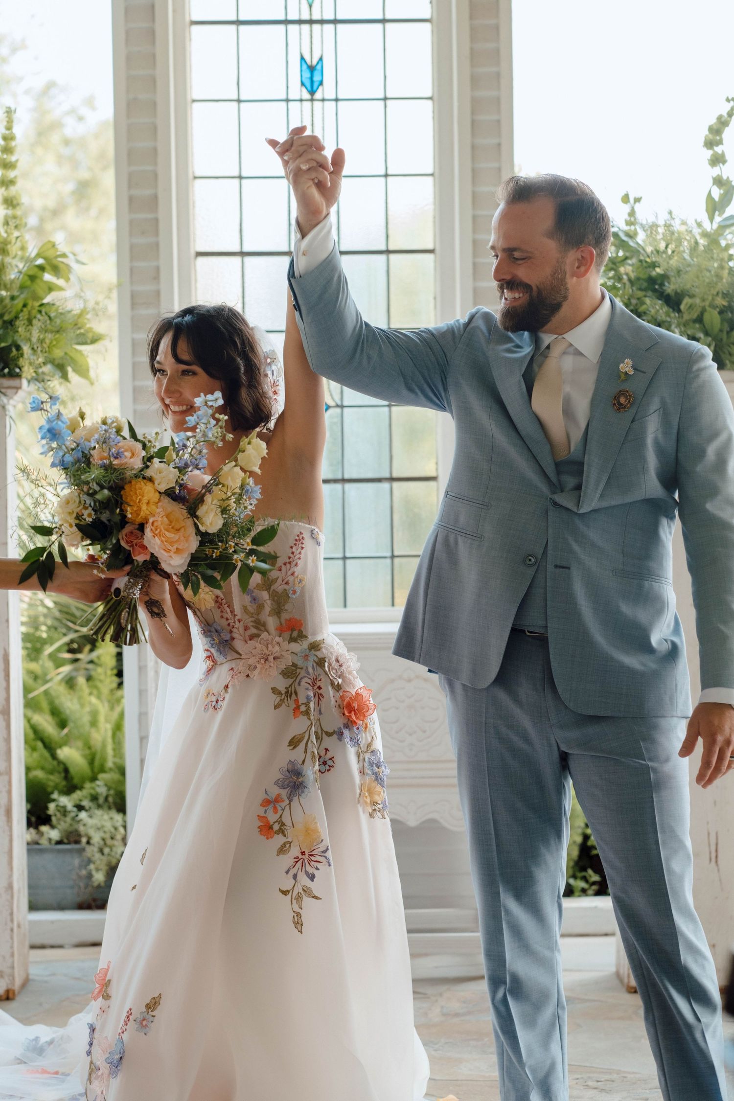 Smiling groom in grey suit raises bride's hand as she holds colorful floral bouquet.