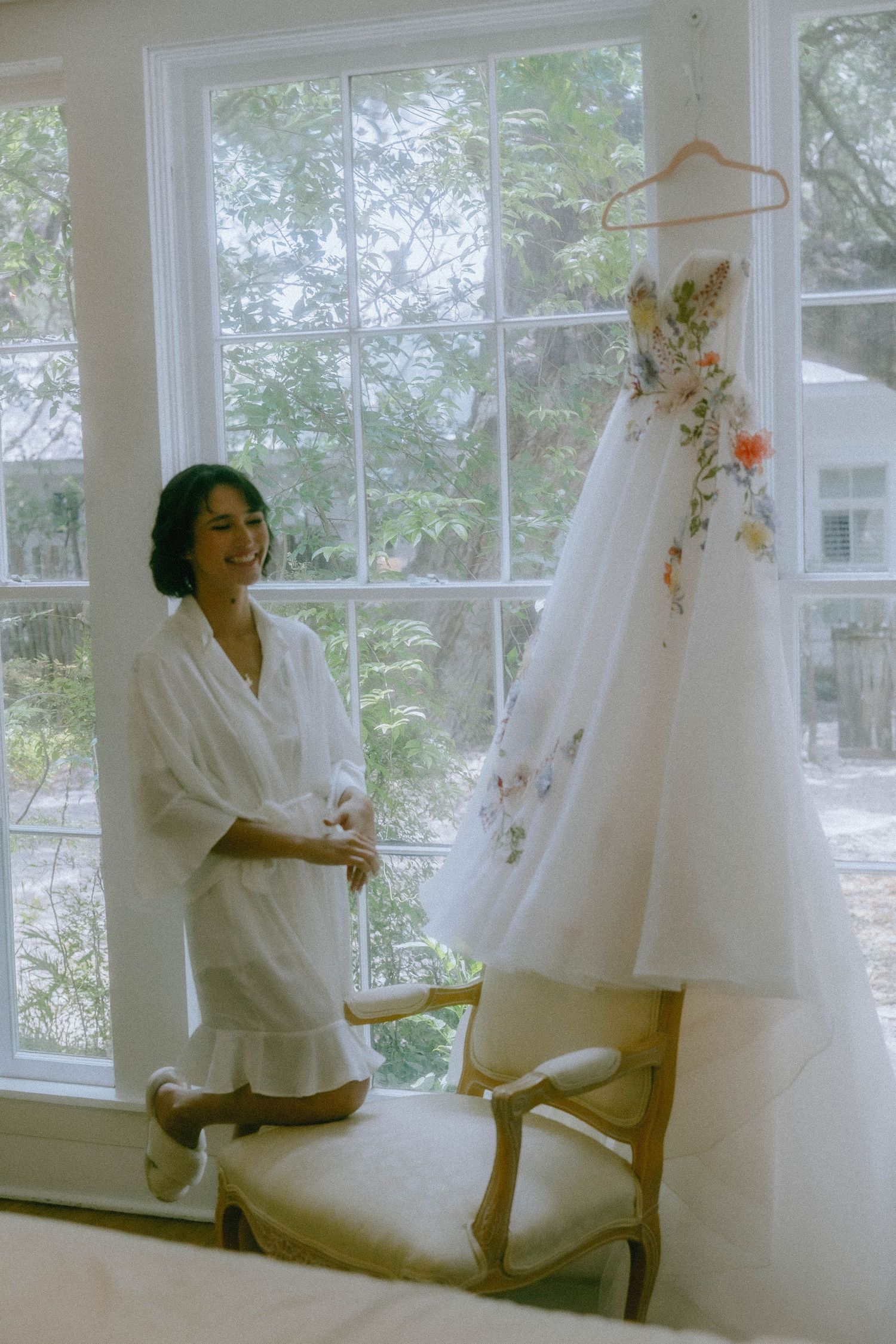 Smiling bride in white robe admires floral embroidered wedding gown hanging by sunlit window.