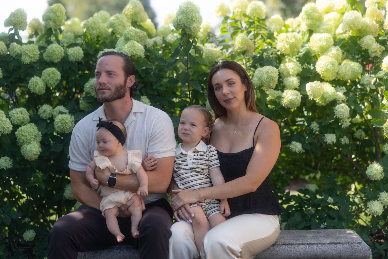 Family of four posing outdoors with lush white hydrangea bushes, parents holding two young children on a sunny day.
