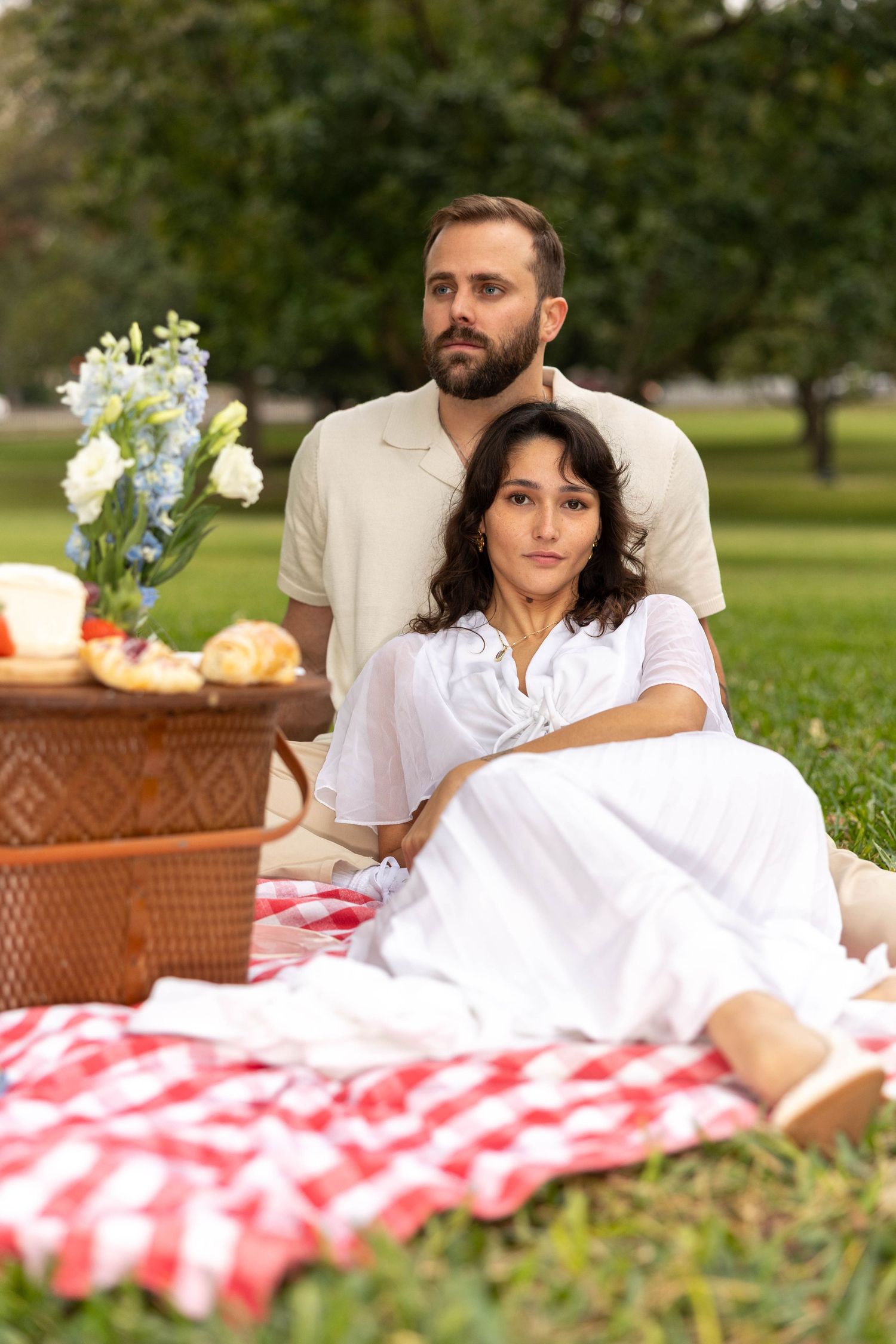 Couple in white outfits poses together on red checkered picnic blanket in green park with wicker basket.