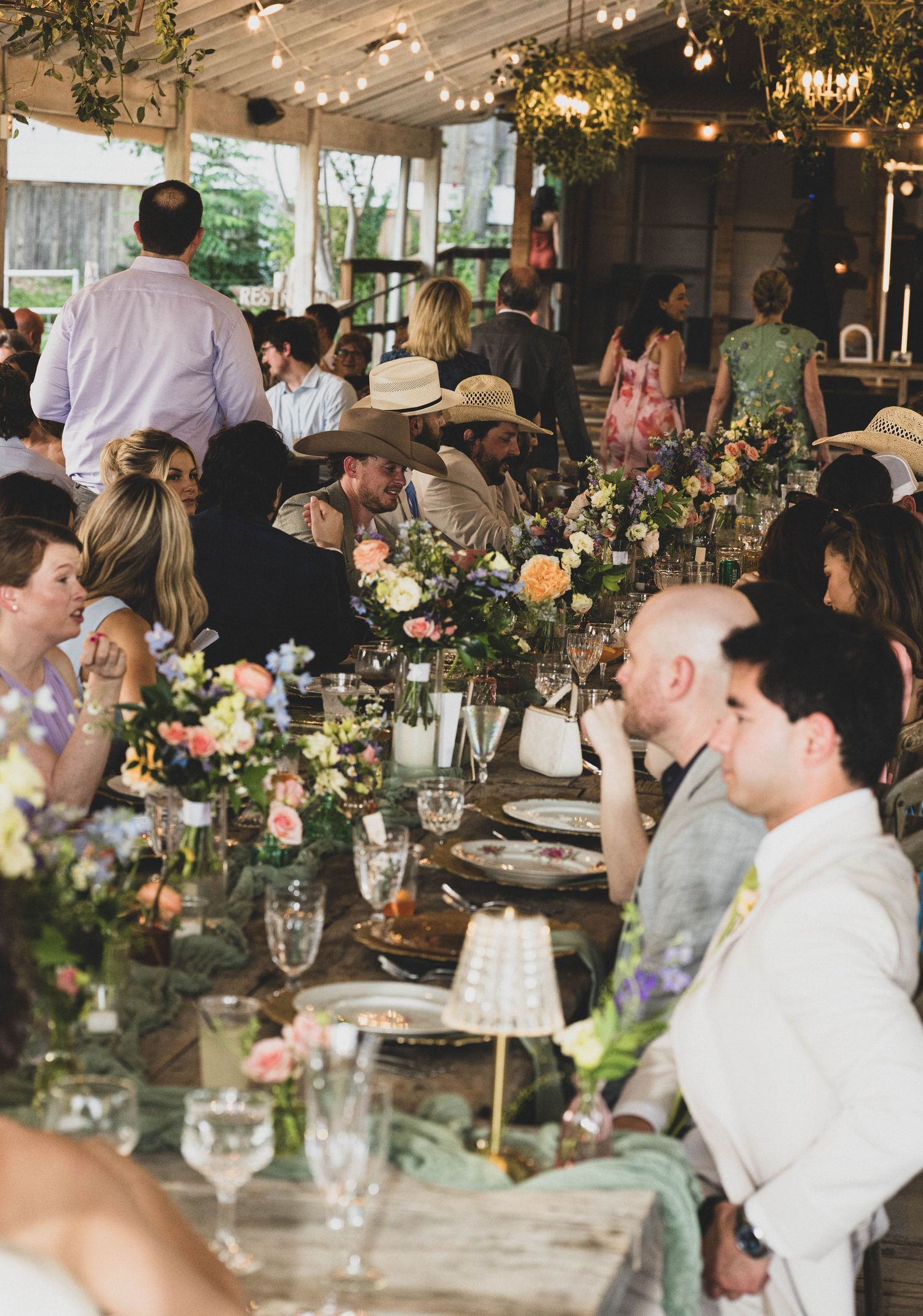 Guests enjoy a lively outdoor wedding reception dinner under string lights with floral centerpieces.