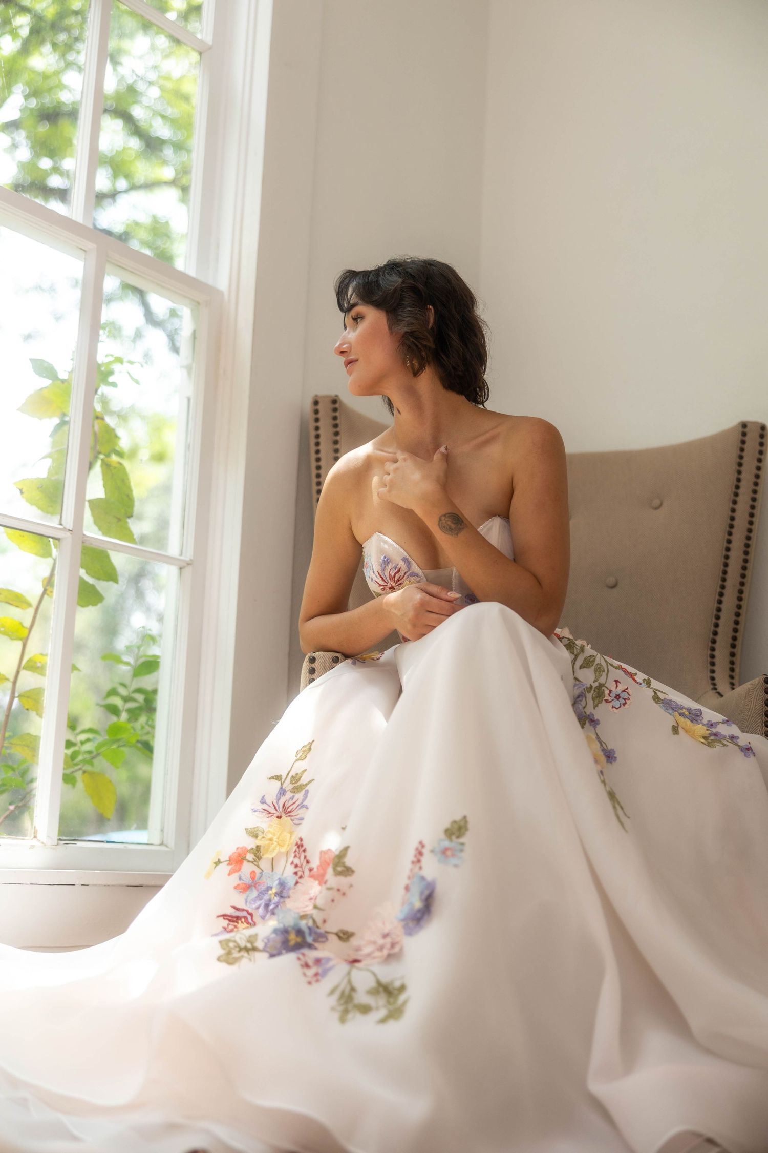 Smiling bride in strapless floral embroidered white ballgown sits by a sunlit window with lush greenery outside.