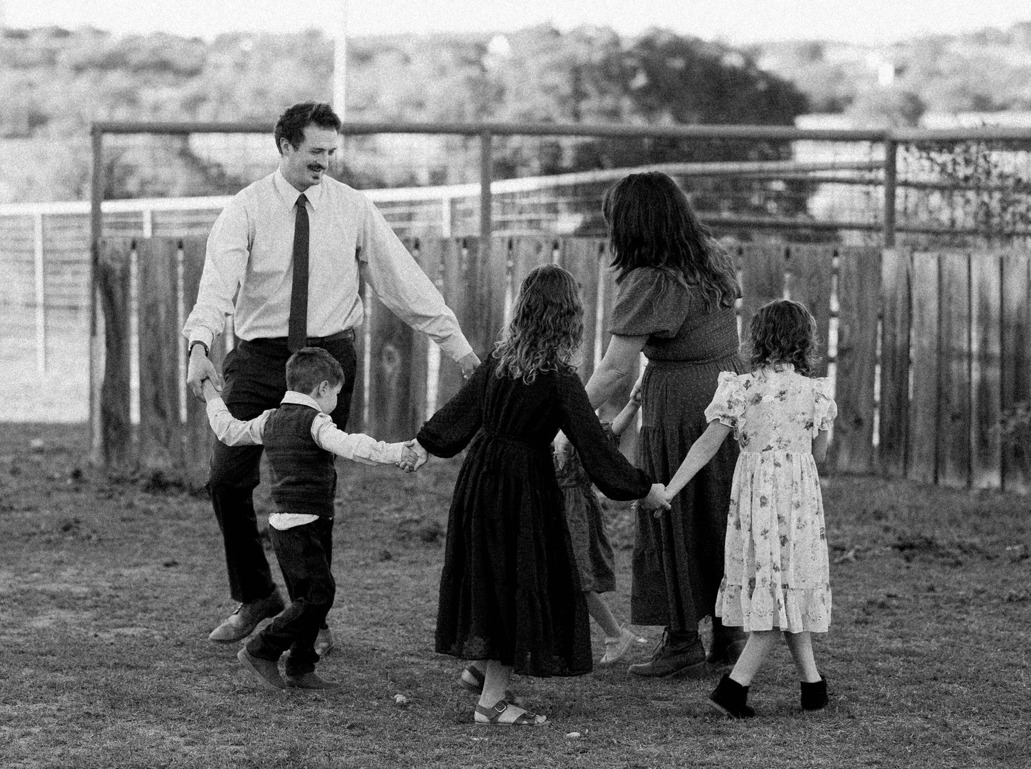 Black and white photo of family of five holding hands in a circle, playing together in an open field near a fence.