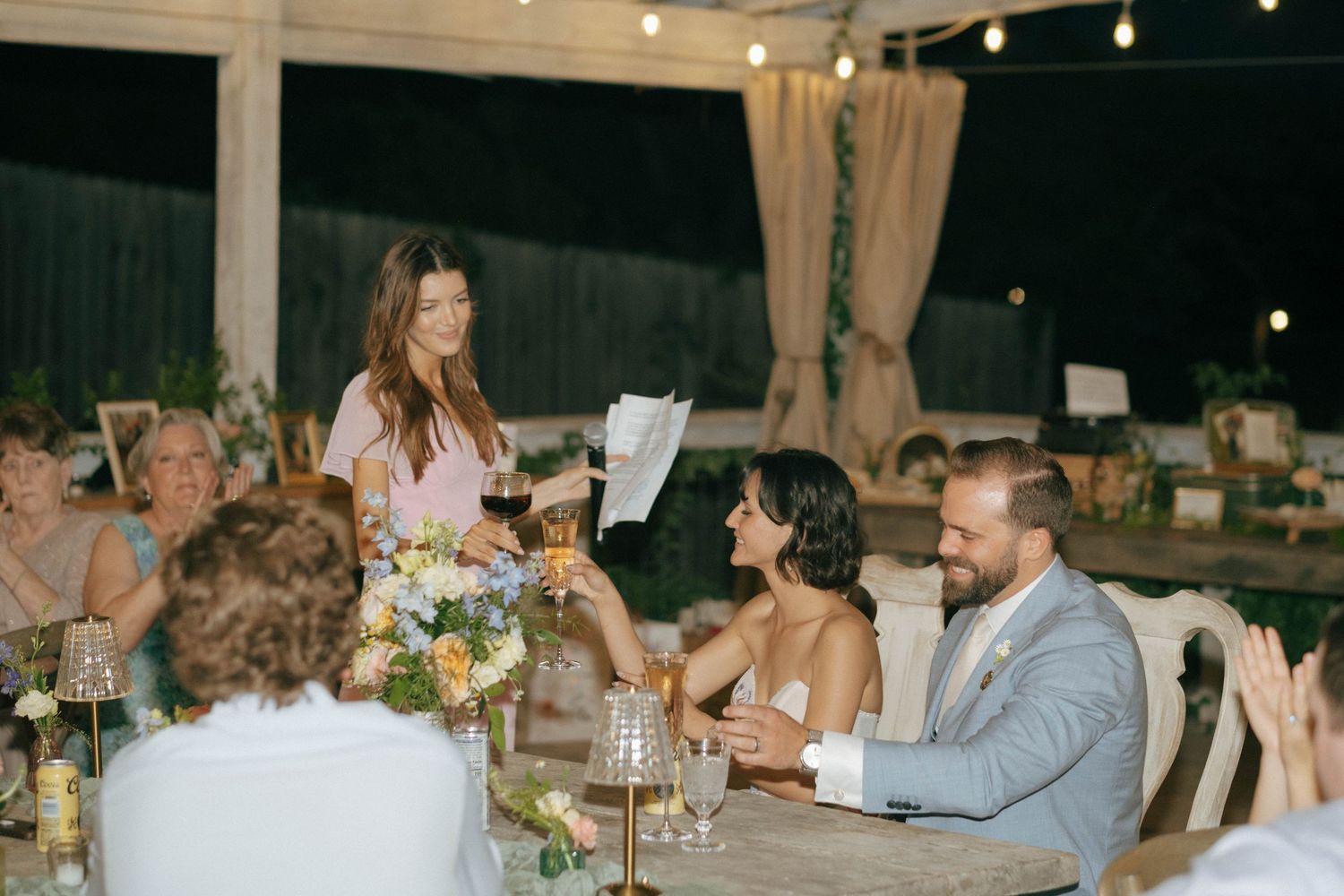 Wedding guest giving a toast speech as the smiling bride and groom listen at an outdoor evening reception dinner.