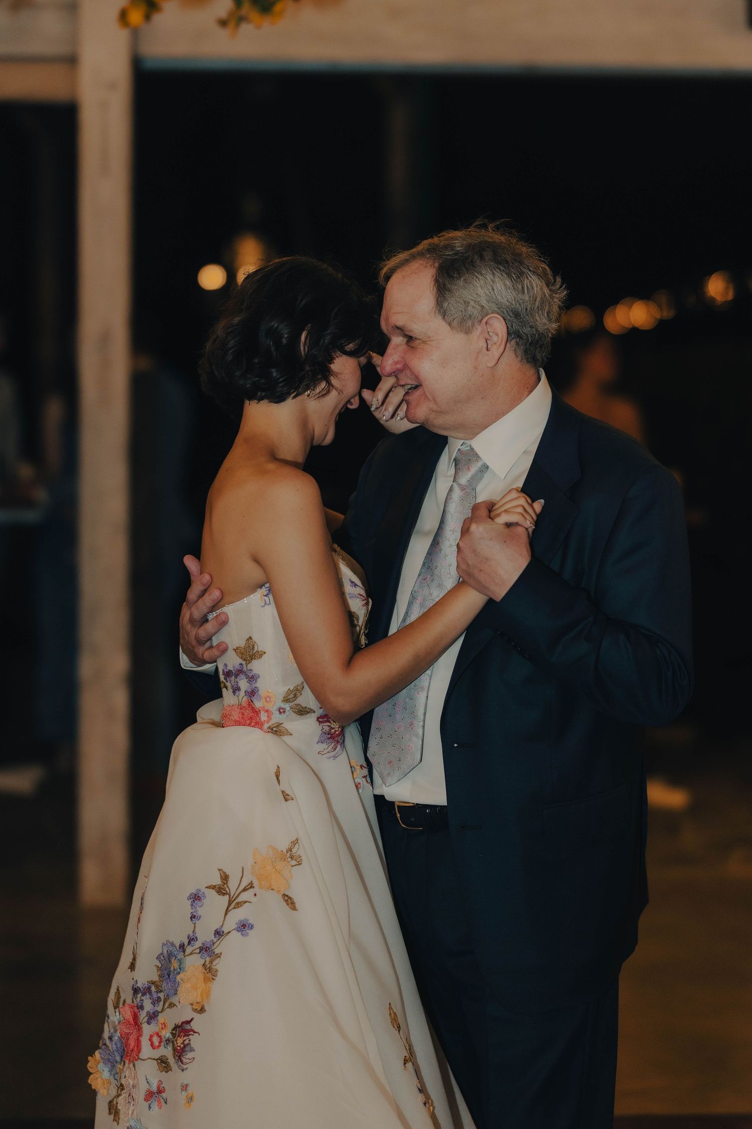 Bride in colorful floral wedding gown shares intimate father-daughter dance at evening reception.