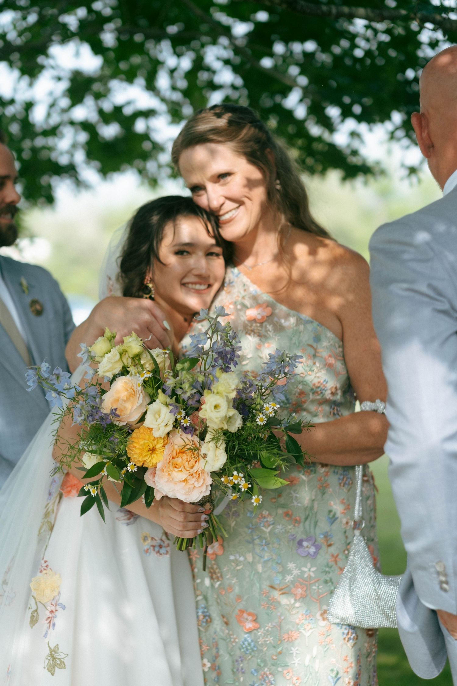 Bride hugs smiling woman in floral dress while holding a colorful wedding bouquet.