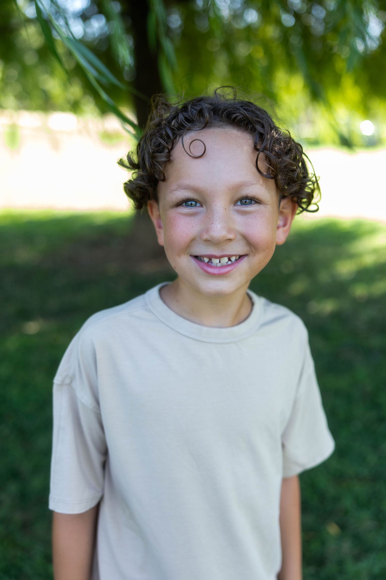 Smiling boy with curly hair and blue eyes wearing a beige t-shirt outdoors in a green park.