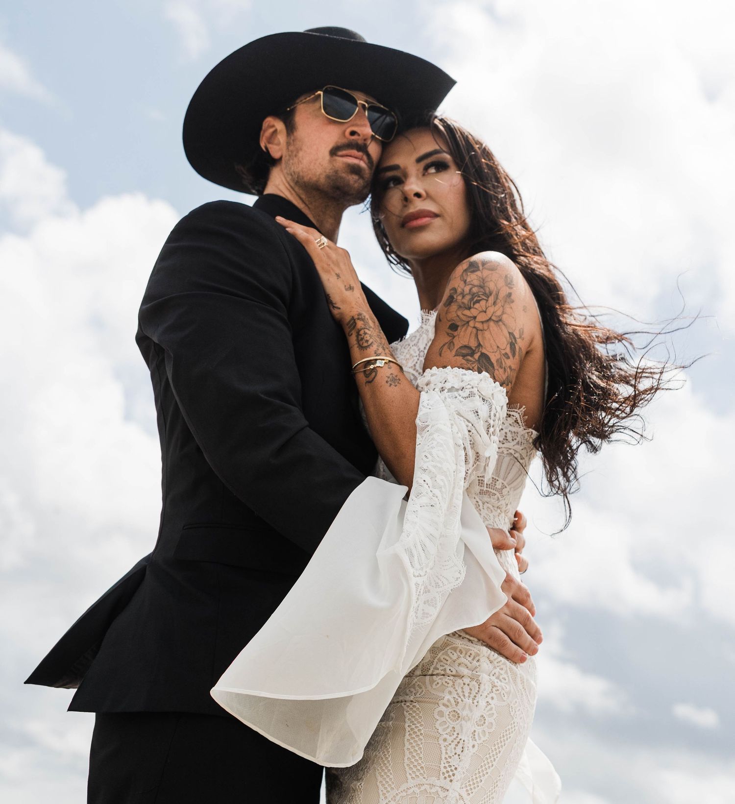 A couple in wedding attire embrace against a cloudy sky background in a romantic outdoor setting.