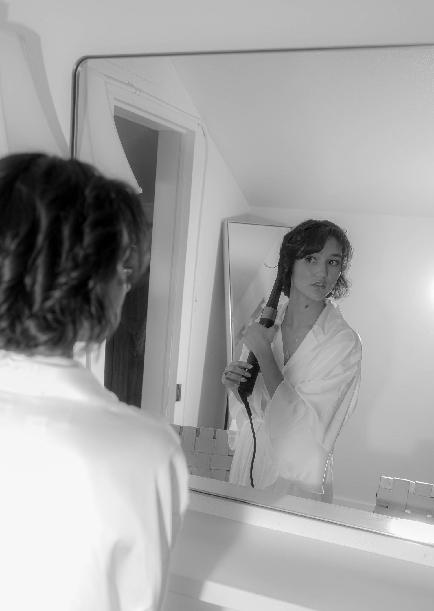 Black and white photo of bride curling hair while looking in mirror before wedding.