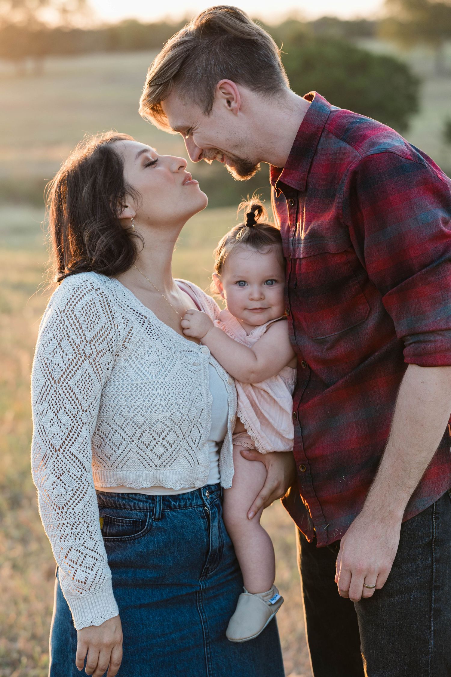 Young couple in field sharing romantic moment while holding their smiling baby at sunset.