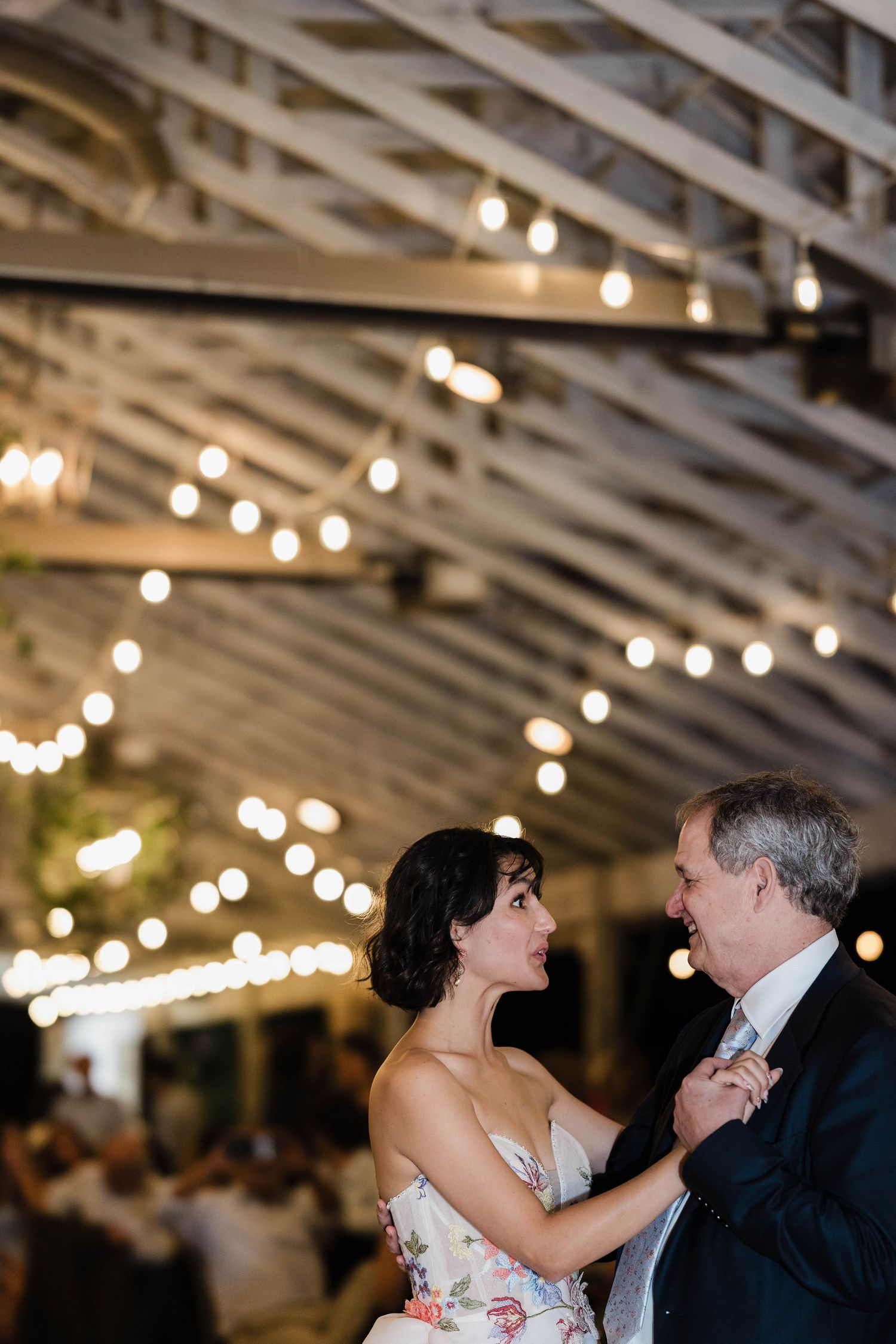 Bride and older gentleman share a tender dance under string lights at a warmly lit wedding reception.