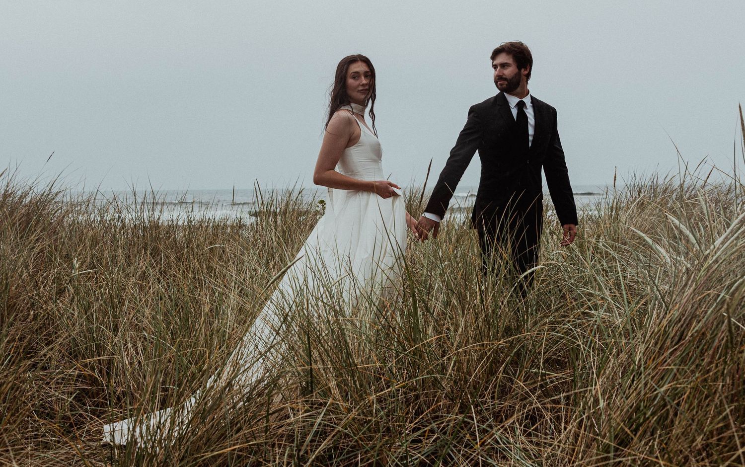 Bride and groom walk hand in hand through tall beach grass, dressed in elegant white gown and black suit.