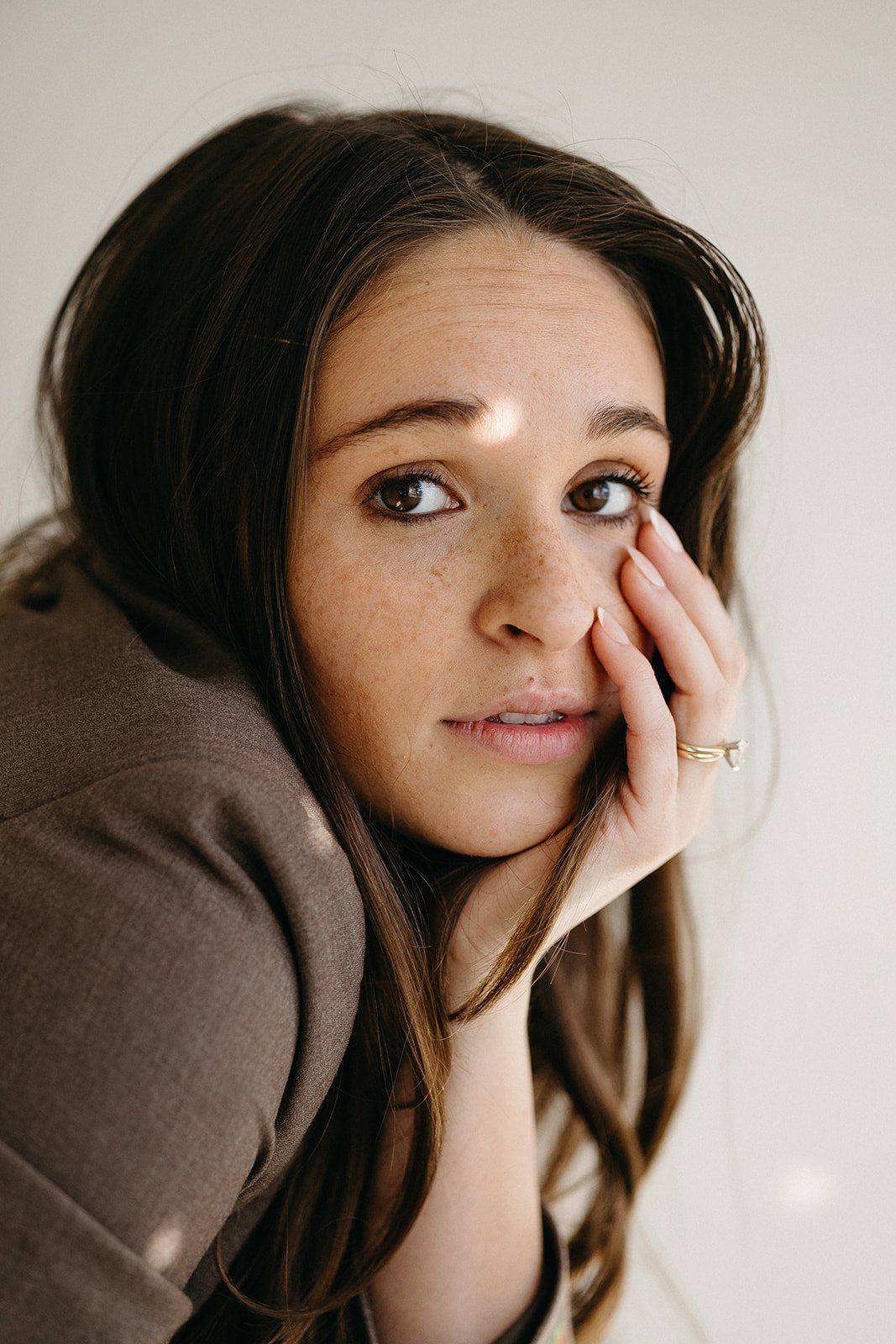 A young woman with freckles and brown eyes rests her hand on her cheek in soft natural light.