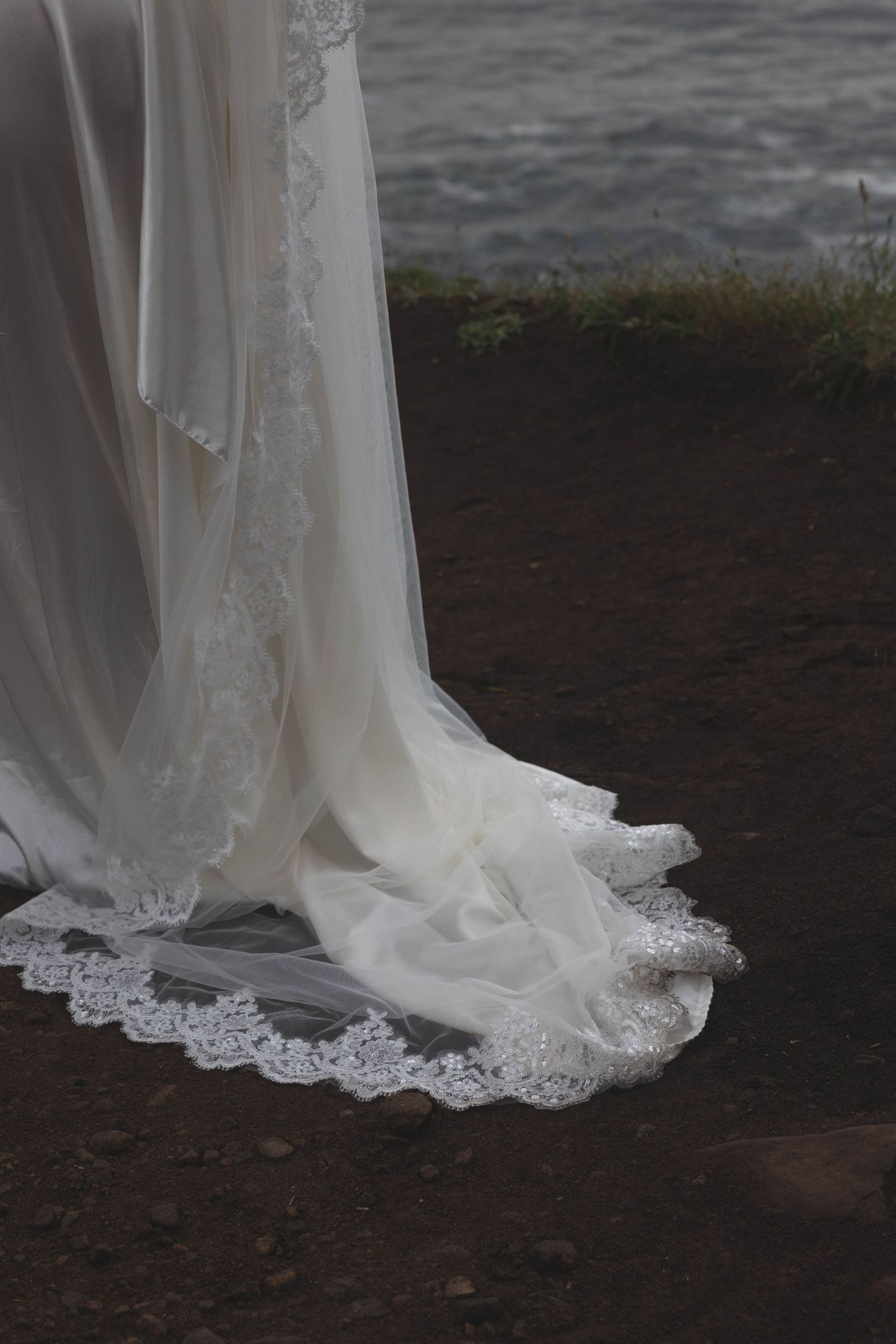 Close-up of a white lace wedding dress train flowing elegantly near a rocky shoreline with water.
