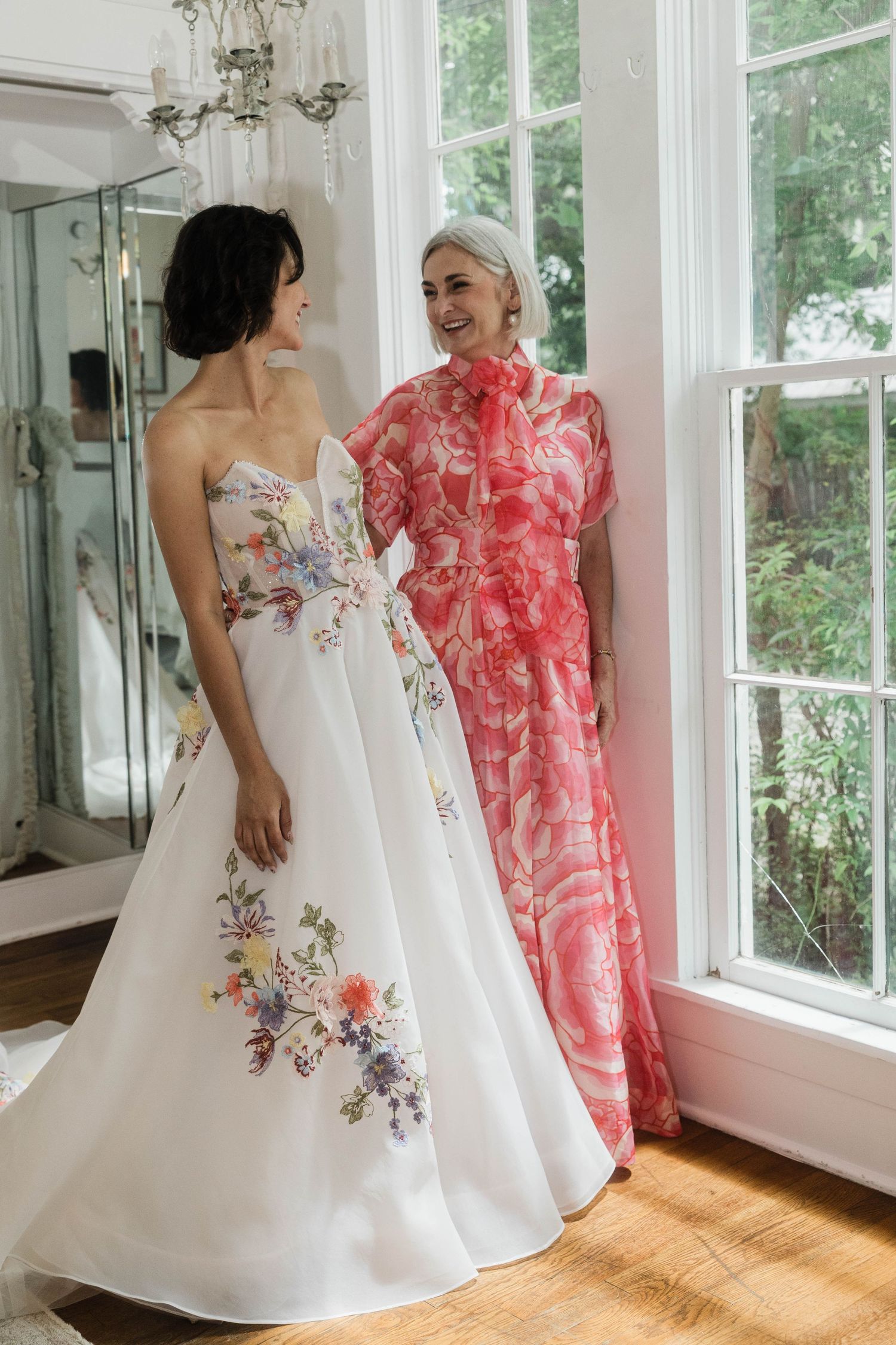 Bride in a floral embroidered strapless gown standing with a smiling woman in a pink dress by a window.