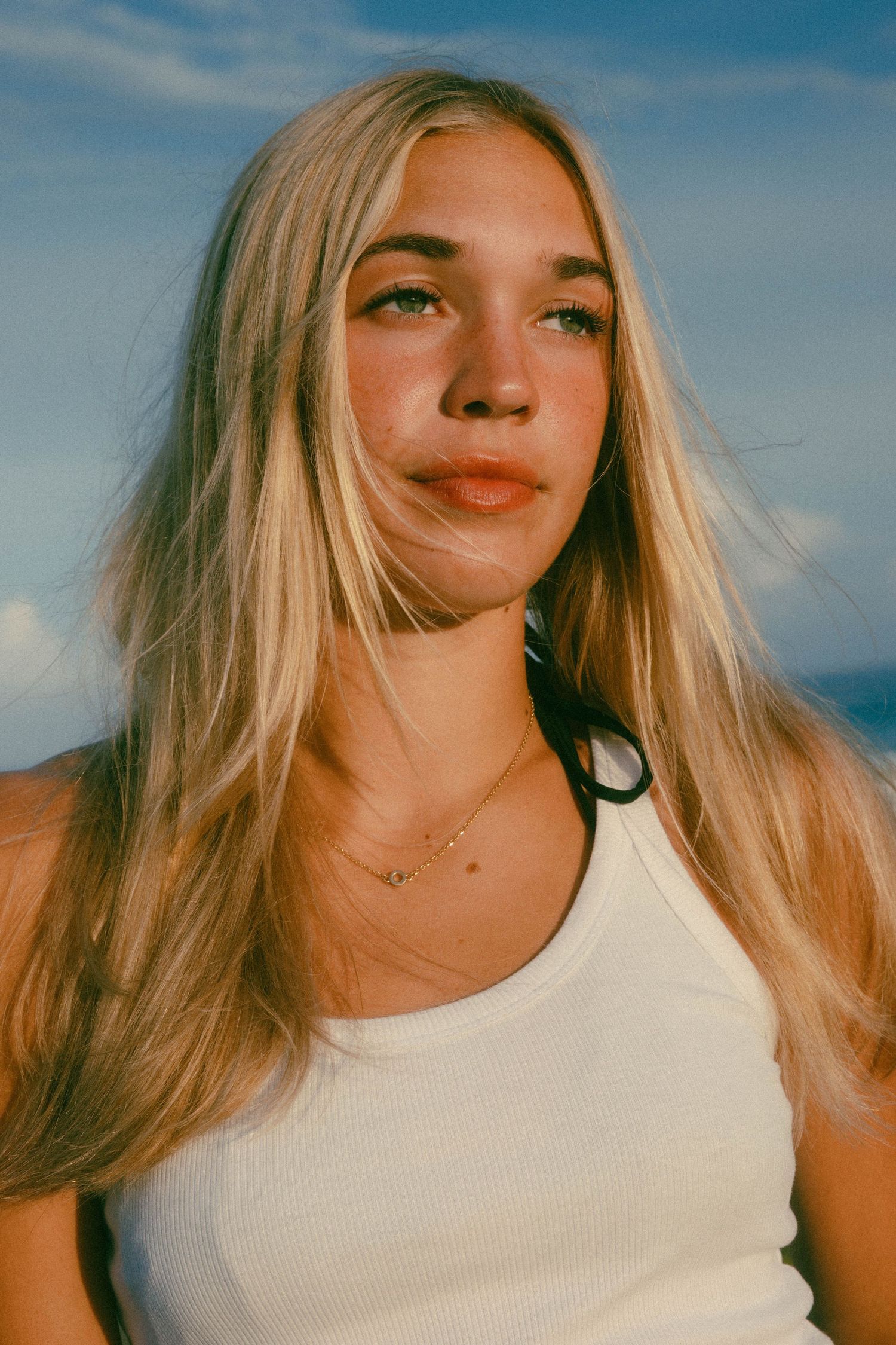 Young blonde woman in white tank top poses outdoors against blue sky with wind in her hair.
